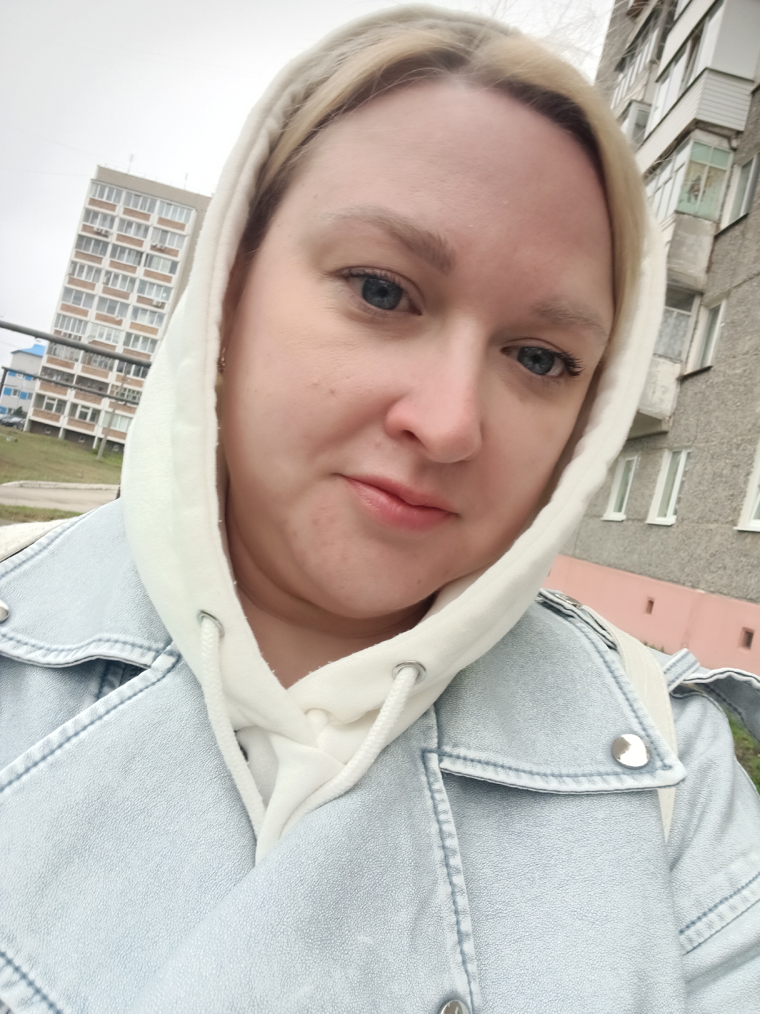 A woman with blonde hair and blue eyes is centered in a selfie-style photograph, wearing a white hooded sweatshirt under a light blue denim jacket. Her face is softly lit, with a slight smile. The background includes a tall apartment building on the left and another grey building on the right. The location appears to be an urban area in Chaikovsky, Russia.