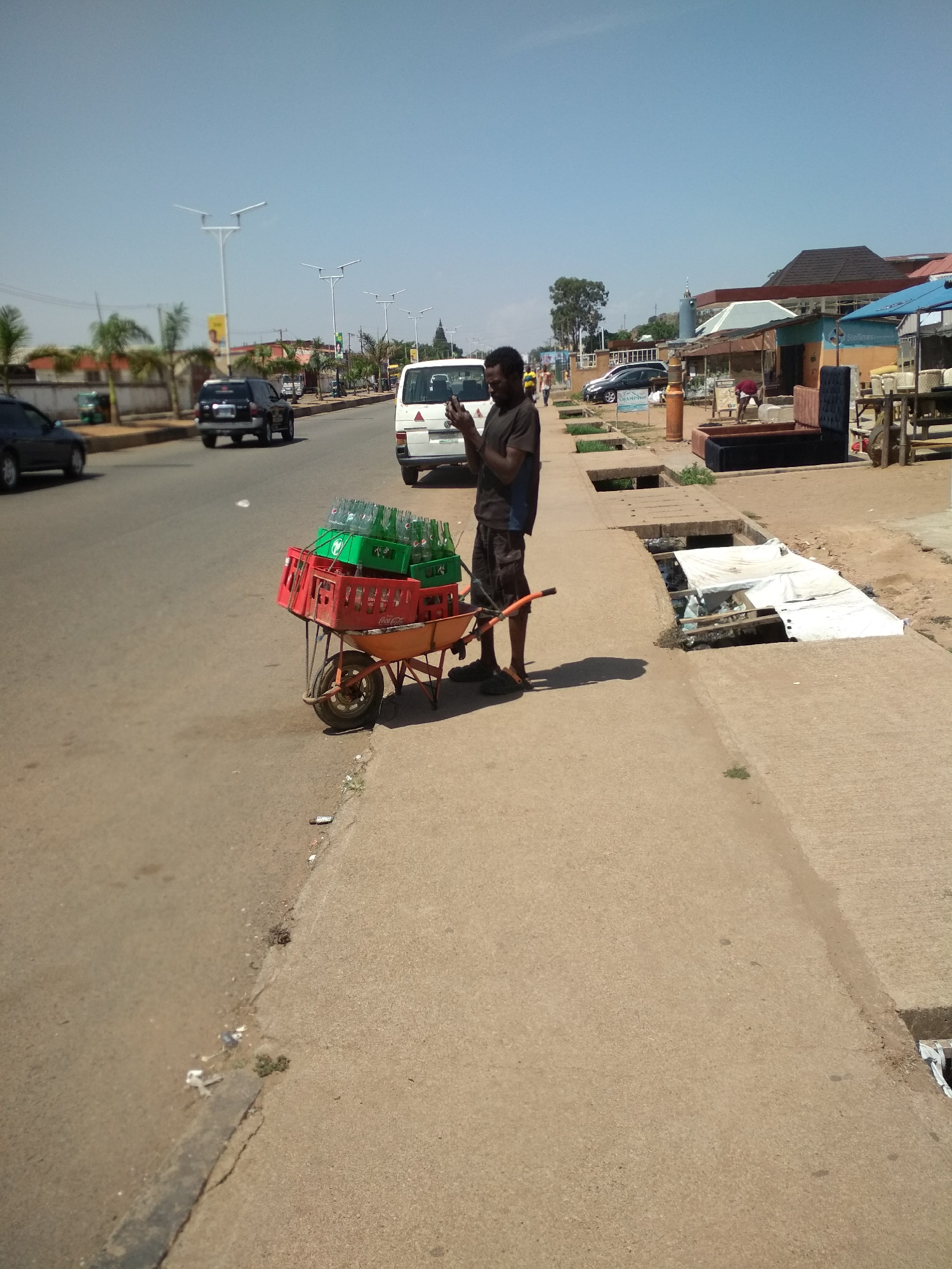 A medium-wide shot depicts a man standing on a sidewalk in Barakin Lerawi, Nigeria. He is holding the handles of a wheelbarrow loaded with stacked crates of green and red bottles. He is facing the camera and appears to be looking at his cell phone. The sidewalk is directly adjacent to a paved road with vehicular traffic. Commercial buildings and street lights are visible along the opposite side of the street. A partially covered open drainage ditch runs along the edge of the sidewalk near where the man is standing. The sky is blue and clear.
