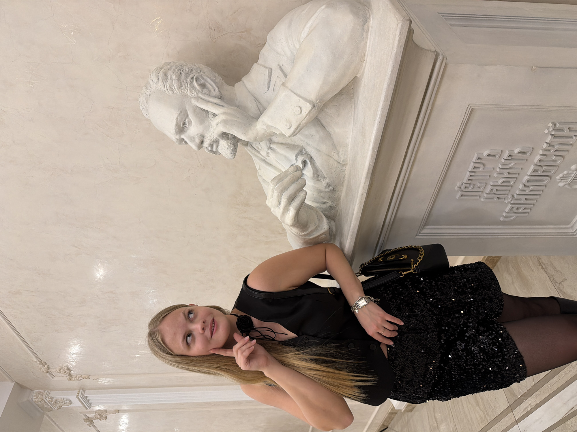 A young woman with blonde hair stands next to a white bust sculpture in what appears to be an interior space. The woman is wearing a black outfit consisting of a vest, a sequined skirt, and dark tights. She has a black purse with a gold chain over her shoulder and is wearing a watch on her wrist. Her right hand is raised to her face, as if in thought or mimicry of the bust.
The bust depicts a man with a beard, wearing a jacket, and resting his head on his hand in a pensive pose. The bust is mounted on a decorated white base with inscriptions in Cyrillic.
Based on visual analysis of the inscriptions and architectural style, the location is possibly the Tchaikovsky Opera and Ballet Theater in Izhevsk, Russia.