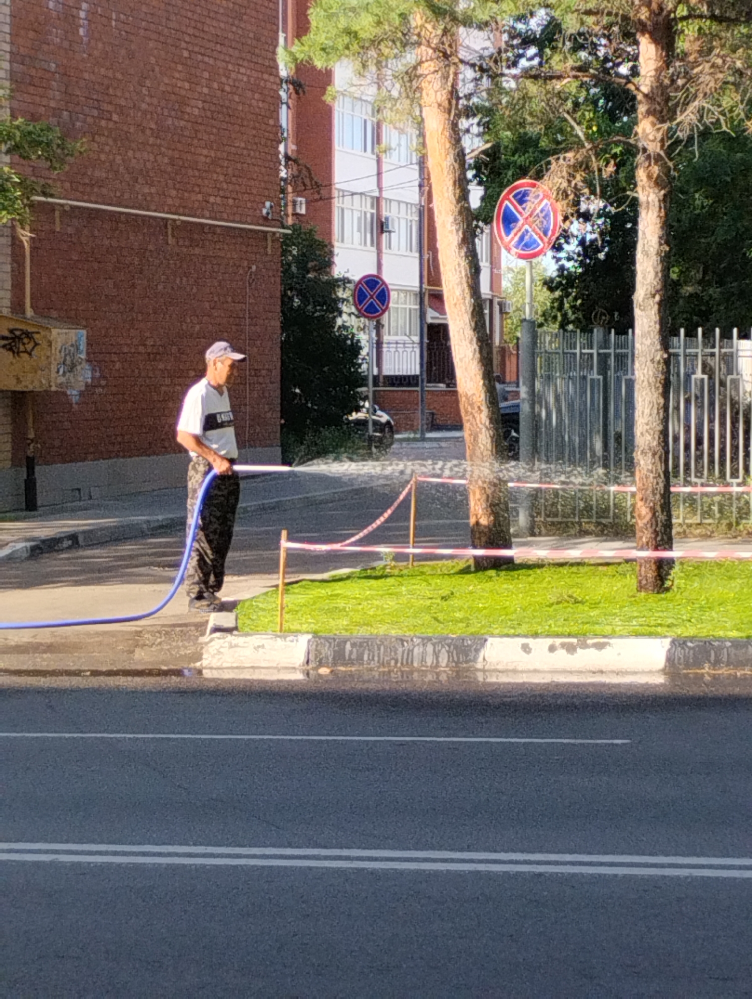 A man wearing a cap, a white t-shirt with a black stripe, and camouflage pants is watering a patch of grass with a blue hose. He is standing on a sidewalk next to a street. Behind him, there is a brick building and a modern building with white balconies. There are two trees in the foreground with red and white caution tape strung between them and poles. Traffic signs indicating "no stopping" are visible behind the trees. The scene appears to be a residential area in the city of Kostanay, Kazakhstan, on a sunny day.
