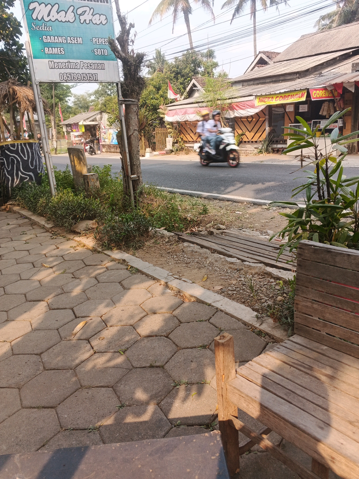 The image shows a street scene in Ungaran, Indonesia, with a focus on a roadside eatery and its surroundings. In the foreground, a hexagonal paver walkway is visible, with a portion of a wooden bench to the right. A tree trunk stands prominently near the left side of the frame, supporting a sign for "Mbak Har" which advertises menu items like "GARANG ASEM," "PEPES," "RAMES," and "SOTO," and provides a phone number. Beyond the walkway and sign, a paved road runs horizontally across the image. On the right side of the road, a building with a corrugated metal roof houses a shop, indicated by banners reading "ANEKA GORENGAN" and "GORENGAN." Two people on a scooter are captured in motion on the road, heading away from the viewer. The environment appears to be a commercial area with trees and some vegetation lining the roadside. The lighting suggests it is daytime, with sunlight casting shadows.