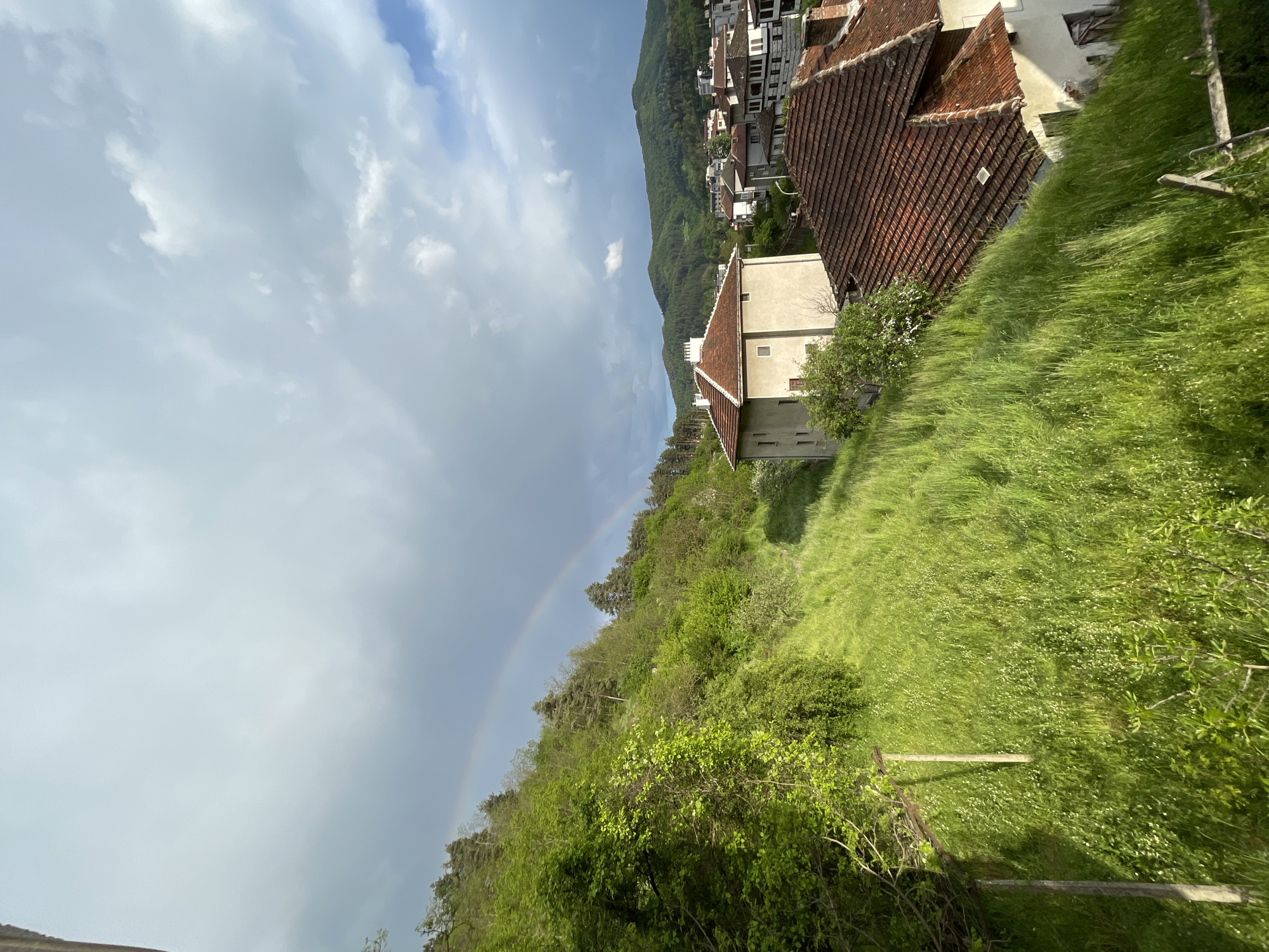 The image depicts a scene in Zlatograd, Bulgaria, featuring a rainbow in the sky. A cluster of buildings with red-tiled roofs sit on a hillside, surrounded by lush green vegetation. The angle suggests the photo was taken from an elevated position. A white building with a tiled roof is also visible amidst the greenery. The foreground is dominated by grass and foliage. The sky is partially cloudy with patches of blue.