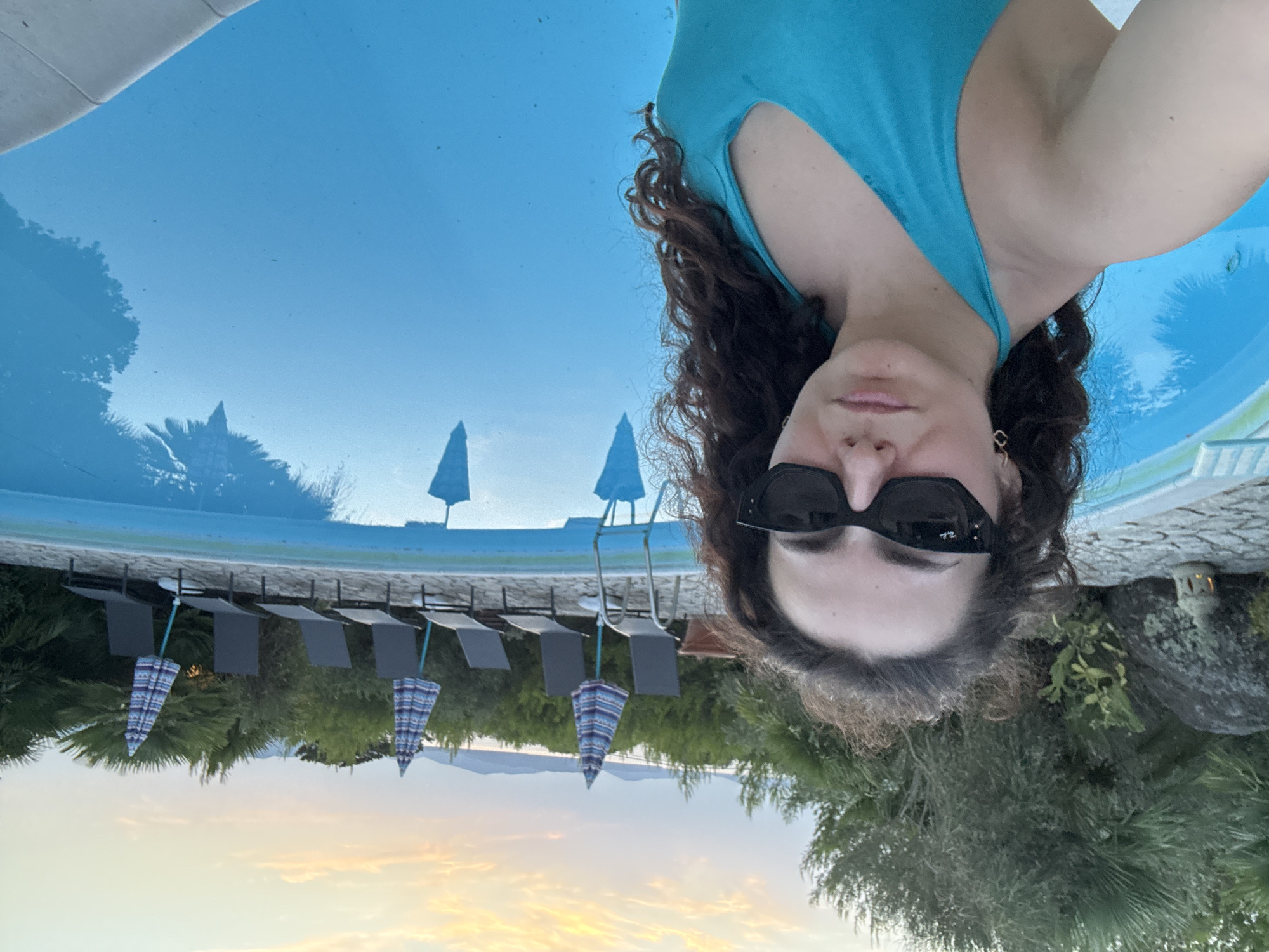 A medium shot captures a woman in a turquoise swimsuit posing for a selfie by a swimming pool at dusk. She is positioned on the left side of the frame, looking directly at the camera with sunglasses on. The pool, filled with bright blue water, occupies the majority of the right side of the image. Beyond the pool, a row of lounge chairs and closed umbrellas are set up, backed by dense green vegetation and trees. In the distance, a body of water is visible, with hills or mountains on the horizon under a pale orange and yellow sky. The overall scene suggests a serene outdoor setting, likely at a resort or private residence, possibly in the Alghero area given the coastal landscape and Mediterranean flora.