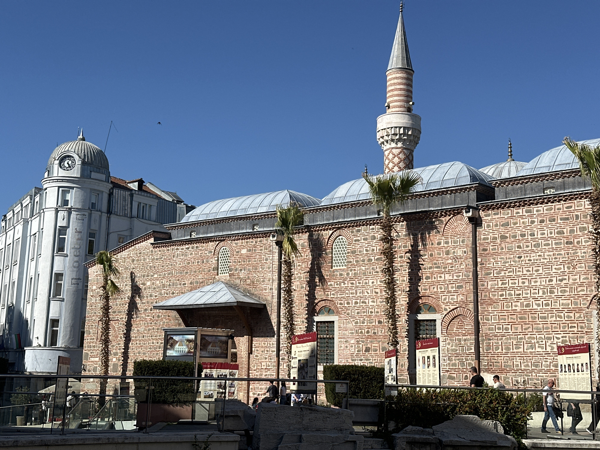 The image shows a street scene in Plovdiv, Bulgaria, featuring the Dzhumaya Mosque. The mosque, constructed of patterned red brick, has a lead-colored roof and a tall minaret with a conical spire. A white building with a clock tower stands next to the mosque. Several palm trees line the front of the mosque, and there are information boards and hedges visible. People are walking by, and there are stone blocks in the foreground. The sky is blue and clear.