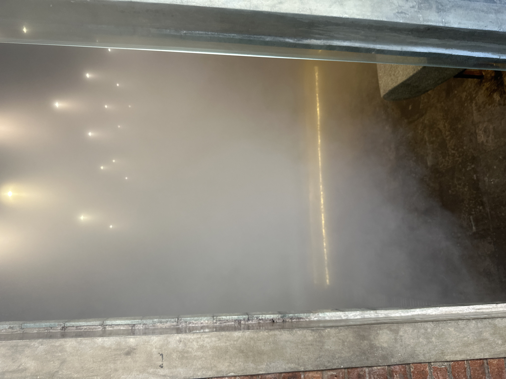 An interior view of a dimly lit space characterized by heavy fog or mist. Numerous small, bright lights are visible suspended in the upper area, diffused by the mist. A horizontal line of yellow light is present at mid-height. In the lower right corner, a portion of a stone bench is visible. The foreground shows a rough concrete or brick wall on the left and a metal pillar or frame on the right. The flooring appears to be dark and possibly wet. The scene evokes an atmosphere of an art installation or a themed environment. The specific location in North Kuta, Indonesia, cannot be determined from the image alone.