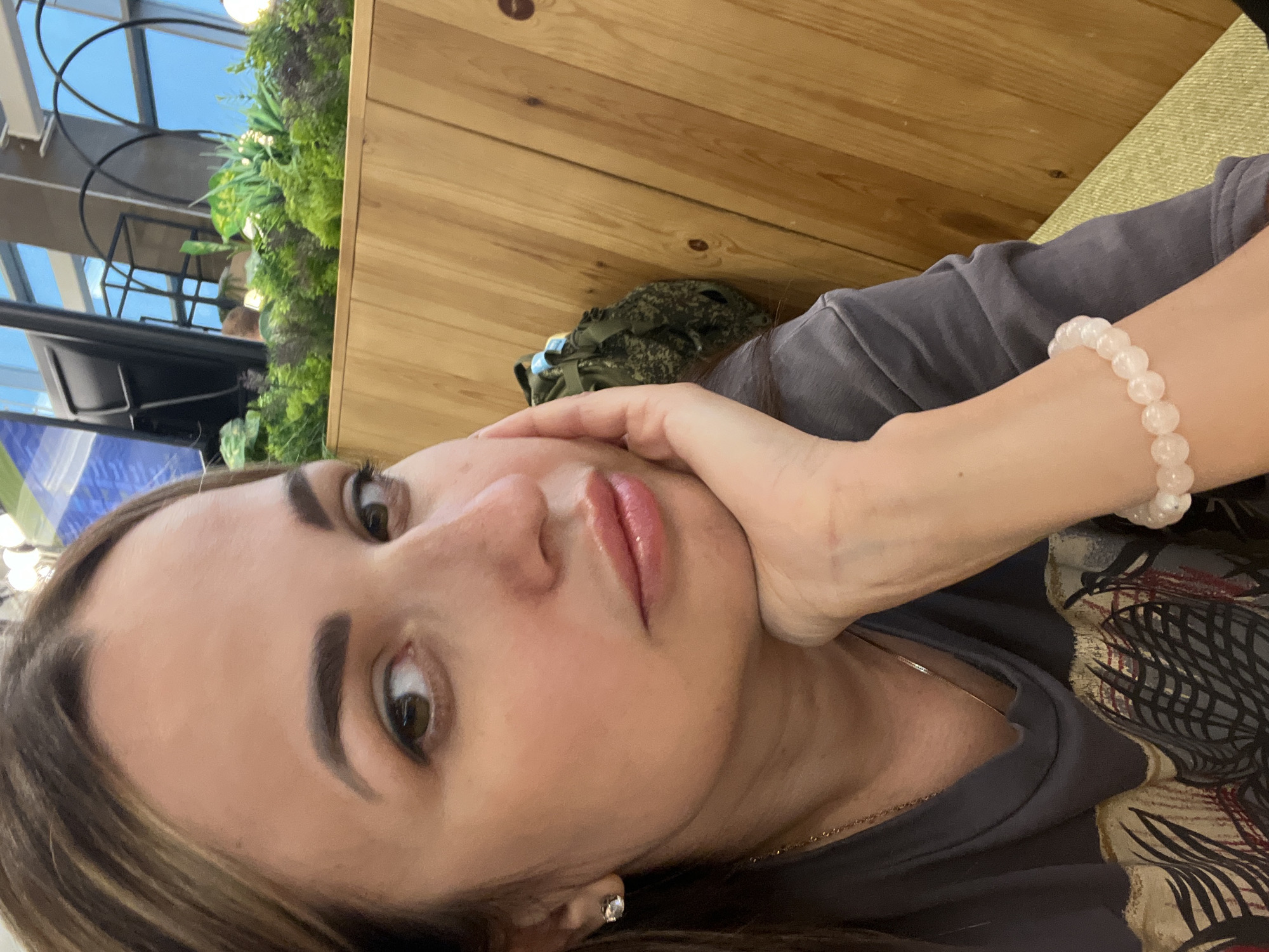 The image is a close-up, waist-up, eye-level selfie of a woman with light brown hair and brown eyes. She is wearing a dark gray t-shirt with a graphic print and a delicate necklace. A bracelet made of light pink beads is visible on her right wrist. Her right hand is resting on her chin, and her left arm is out of frame. She is looking directly at the camera with a neutral expression. The background consists of light-colored wooden paneling and a blurred view of artificial plants and building structures, suggesting an indoor setting. The lighting is soft, and the focus is on the woman's face and upper body.