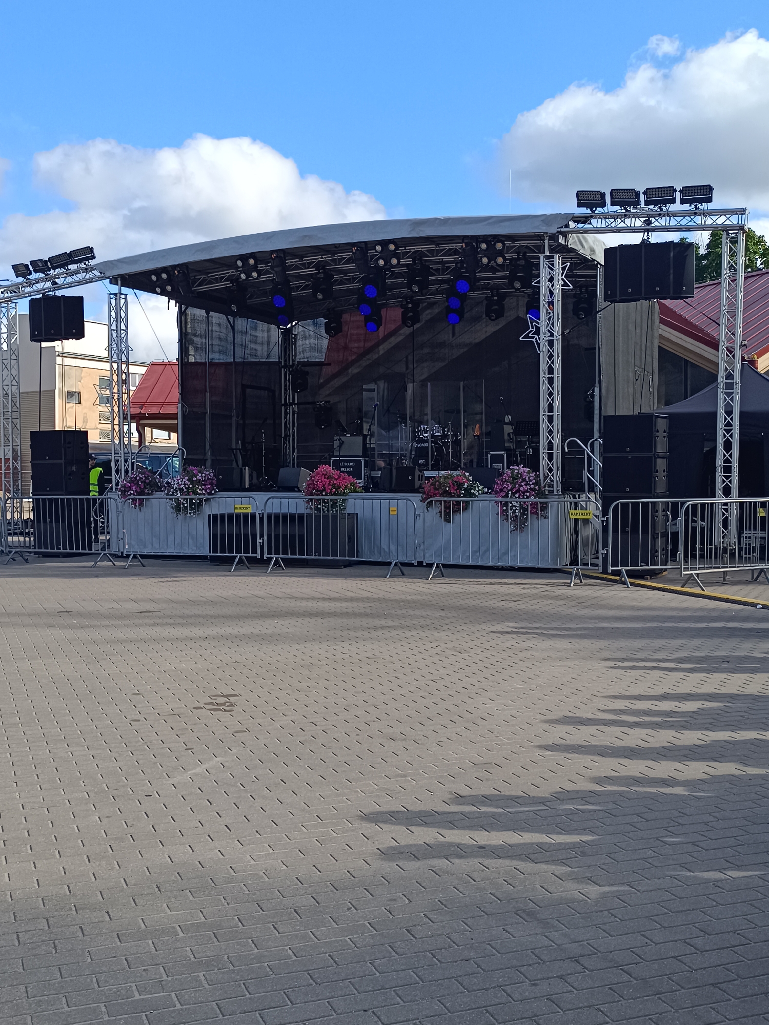 The image depicts an outdoor stage setup in the city of Aizpute, Latvia. The stage is covered with a gray tarp and equipped with professional lighting and sound systems, including large speakers mounted on truss structures. The stage is fronted by a metal barrier, behind which several planters filled with pink and purple flowers are visible. A person wearing a bright yellow vest stands near the stage. The ground in front of the stage is paved with interlocking brick pavers, and shadows from the stage and surrounding objects are cast across the pavement. The sky above is blue with scattered white clouds. A building with a red roof is visible behind the stage to the right.