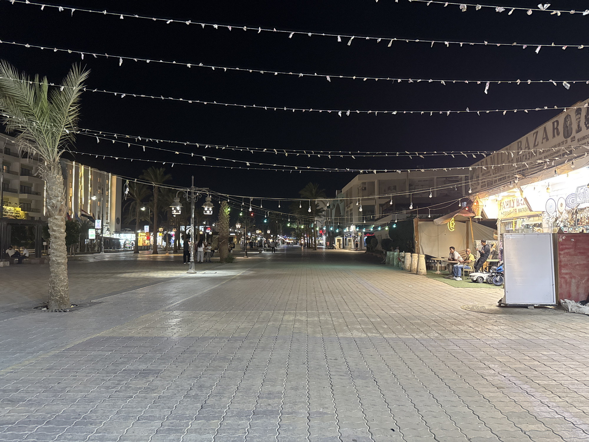 Here is a description of the image:
The image depicts a street scene at night. Overhead, strings of lights crisscross the expanse, illuminating the street below. The street itself is paved with interlocking blocks. On the left side of the image, a palm tree stands tall, adjacent to buildings with illuminated facades. Pedestrians are visible, moving about on the street. On the right side, a bazaar-like structure is present, with individuals seated nearby, creating a sense of activity and commerce.