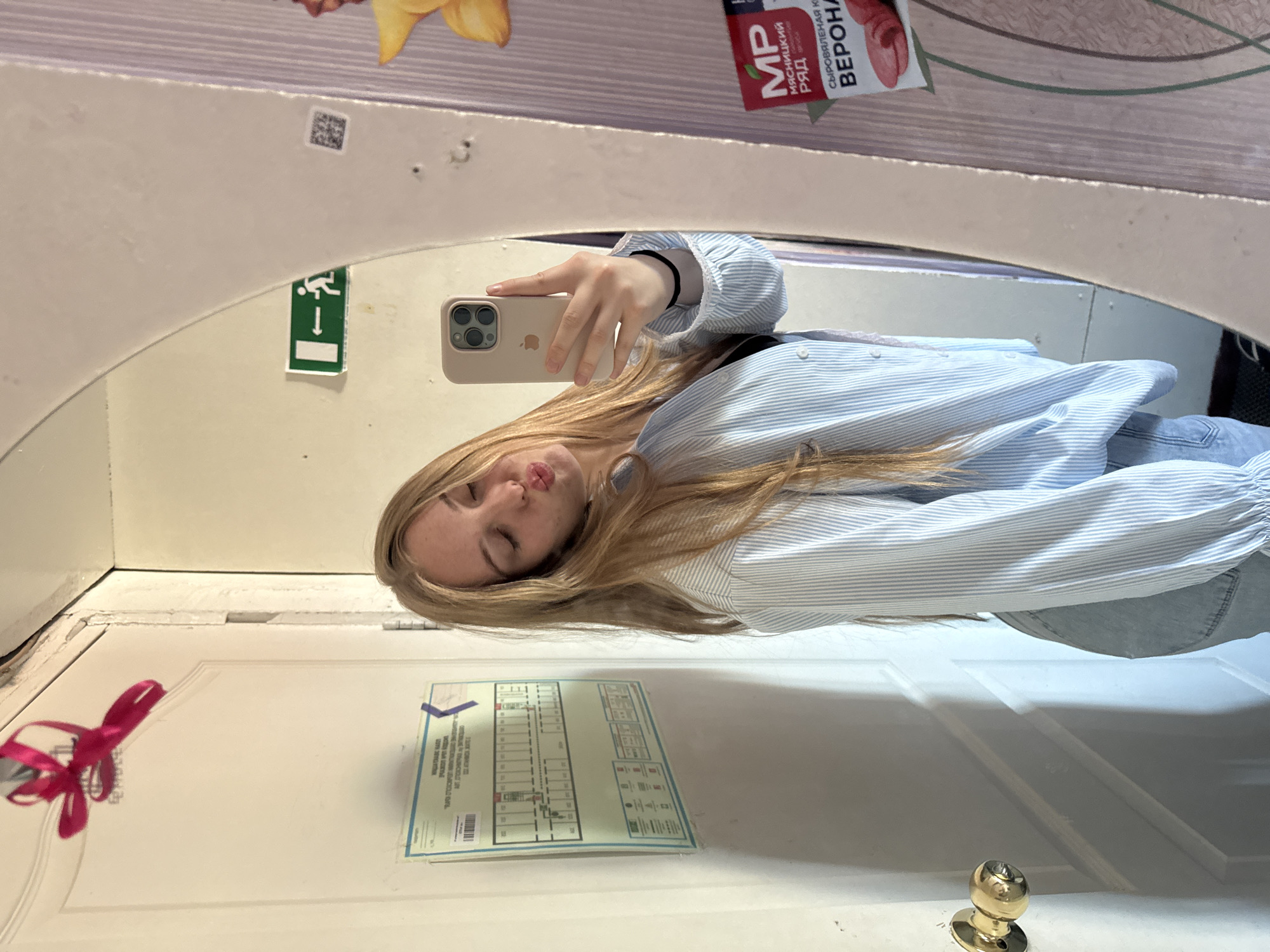 A young woman with long blonde hair is taking a selfie in a mirror. She is wearing a light blue striped shirt and jeans. The mirror is reflecting a train compartment, with a white door decorated with a pink ribbon and a safety evacuation plan. There's an emergency exit sign visible above her head in the mirror. The compartment appears to be in a train, and there's an advertisement in Russian at the top of the mirror, possibly identifying the location as Russia, potentially Perm, Russia based on the product or advertisement.