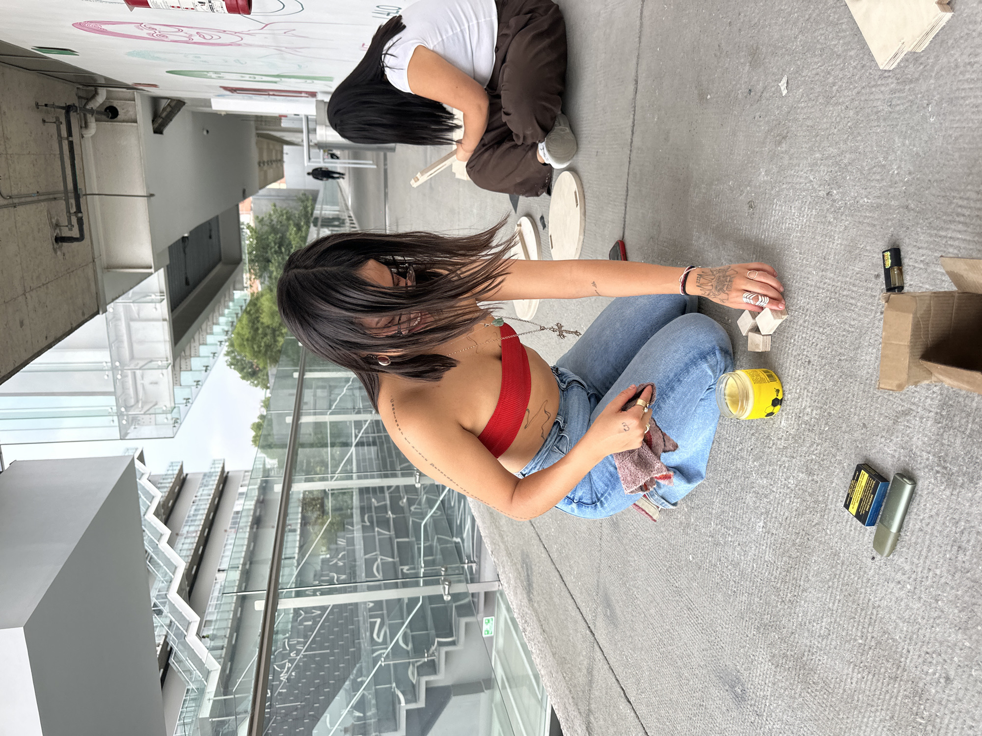 Two women squat on a textured gray surface in Mexico City, Mexico, with modern architecture including glass-walled staircases visible in the background. One woman wears a white shirt and brown pants, working with wooden discs and tools. The other woman wears a red top and blue jeans, interacting with small wooden blocks, a yellow jar, and other items. Tattoos are visible on her hands and arms. A box and a packet are placed to the side. A mural is visible on the white wall behind.