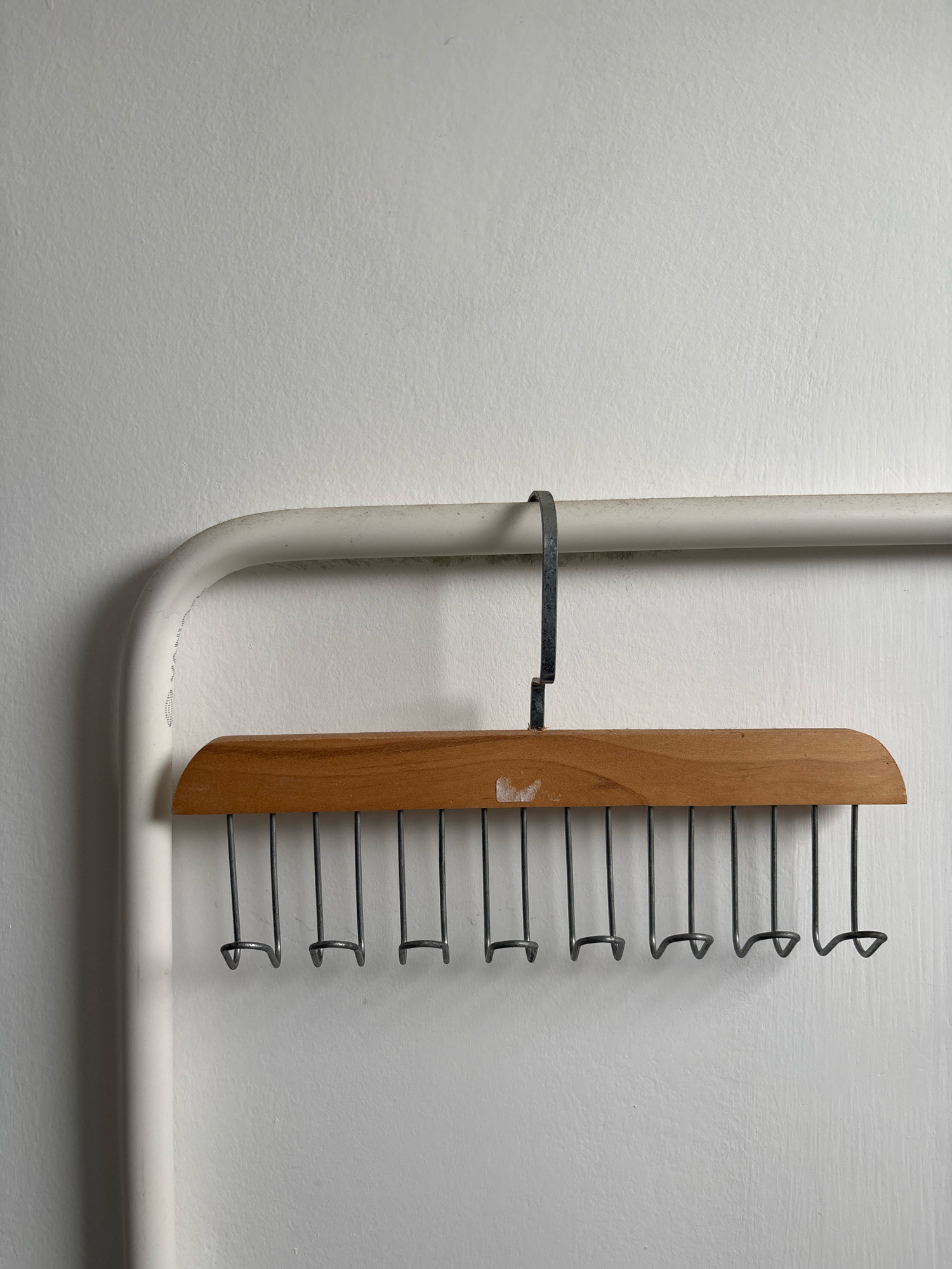 A wooden hanger with numerous metal loops is hanging from a white metal pipe against a textured white wall. The hanger has a slightly curved wooden bar with a darker circular imprint near the center. Below the wooden bar, a series of thin, dark metal wires hang vertically, each ending in a small, U-shaped loop. The pipe runs horizontally across the frame, supported by a vertical section on the left side that curves to meet it. The wall appears to be indoors, with subtle shadows cast by the objects. There are no people or animals present in the image. The scene is static and appears to be a still life. The location of this scene in Nha Trang, Vietnam, is not discernible from the visual information provided.