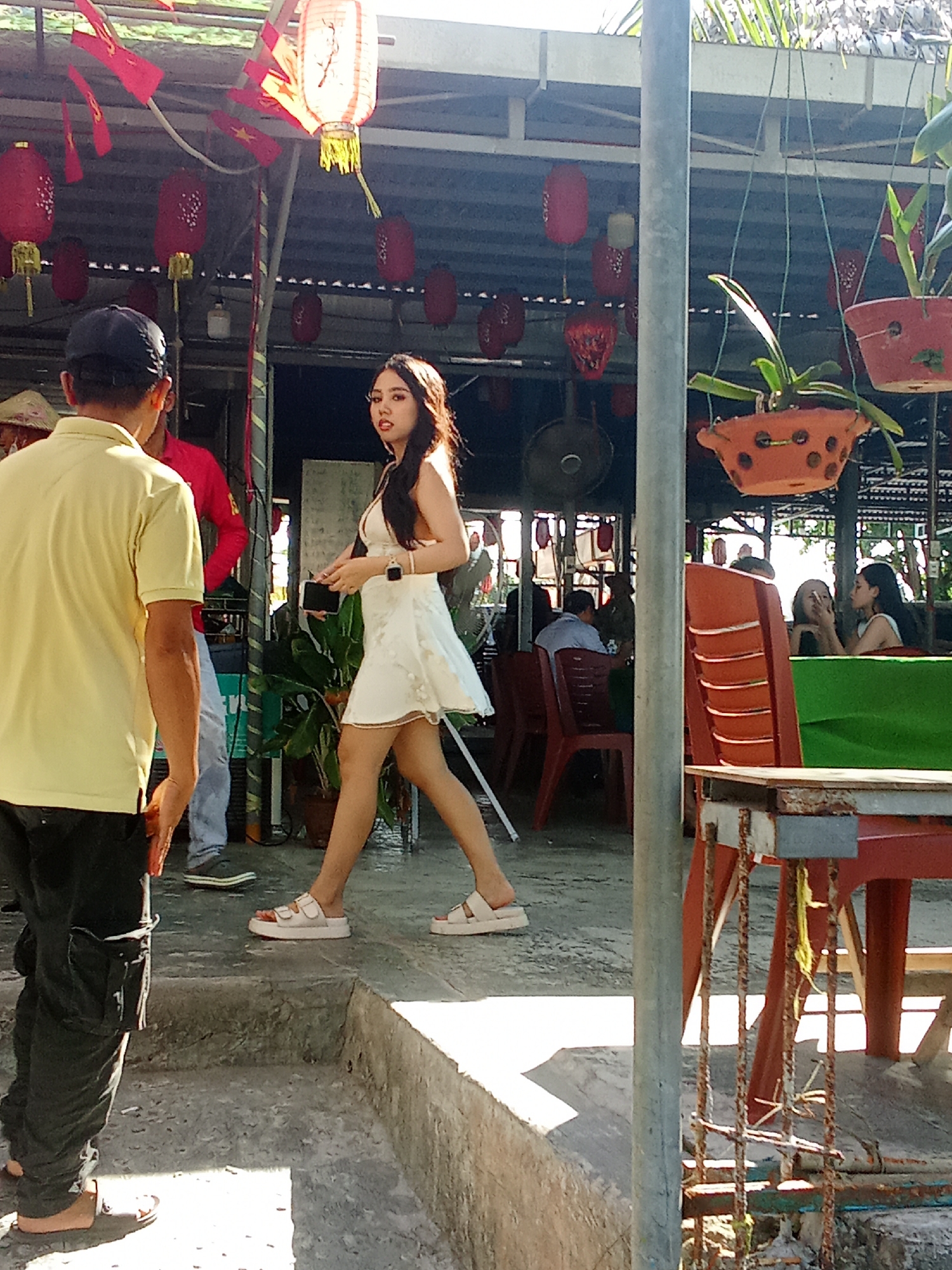 The image depicts a scene outdoors, possibly a cafe or restaurant, with a distinct Vietnamese ambiance, suggested by the presence of red lanterns and tropical foliage. In the foreground, a person wearing a yellow polo shirt and black pants is walking away from the camera. To their right and slightly behind, a young woman in a white dress is walking, looking back towards the camera. She is wearing white platform sandals and jewelry on her wrists.
The background reveals more of the setting, with a covered area supported by pillars, and red chairs and tables arranged for patrons. Several people are seated in the background, some engaged in conversation. Red lanterns are hanging from the ceiling, along with some potted plants. The sunlight suggests it is daytime, and the overall atmosphere appears casual and relaxed. The rough texture of the concrete steps and ground indicates an outdoor, possibly somewhat rustic, environment. The scene likely takes place in Đà Nẵng, Vietnam, given the cultural context implied by the decorations. No visible text is present in the image.