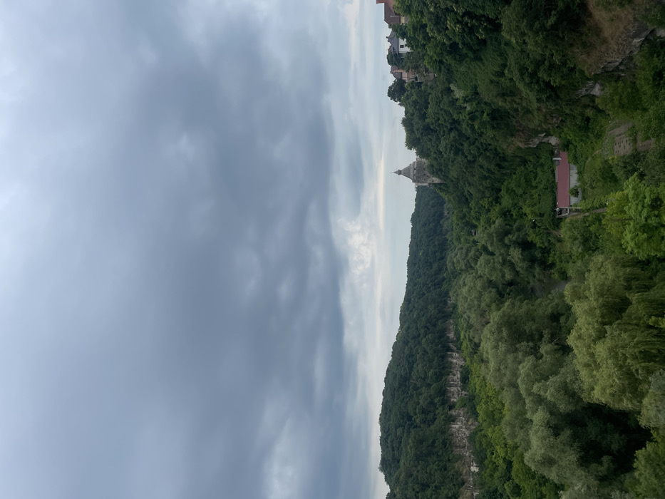A cloudy sky dominates the view above a lush green hillside. Atop the hill, a tower or castle-like structure stands tall. Thick vegetation covers the hillside, with trees forming a dense canopy. Buildings are seen at the upper right corner. The image appears to be taken from a high vantage point, looking across a valley or gorge filled with trees.