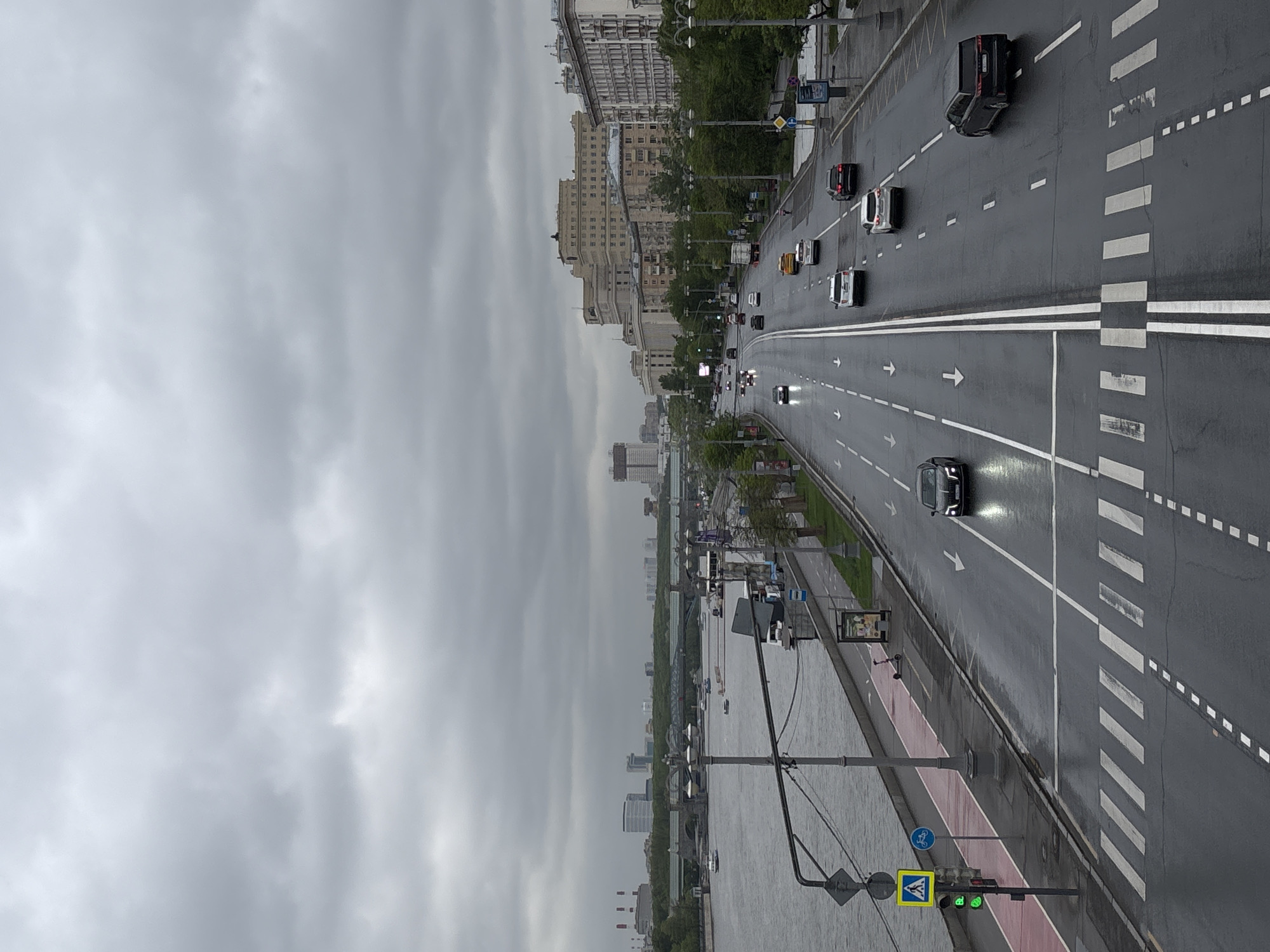 The image shows a view of a wide, wet roadway in Moscow, Russia, with multiple lanes of traffic moving away from the camera's perspective. Cars are visible on the road. Along the roadway's edge, there is a pedestrian walkway with a red stripe, bicycle lane markings, and a pedestrian crossing sign. A traffic light is green. In the background, buildings of varying heights are visible against an overcast, cloudy sky. The roadway appears to be near a body of water, with the embankment and waterway seen on the left side of the frame.