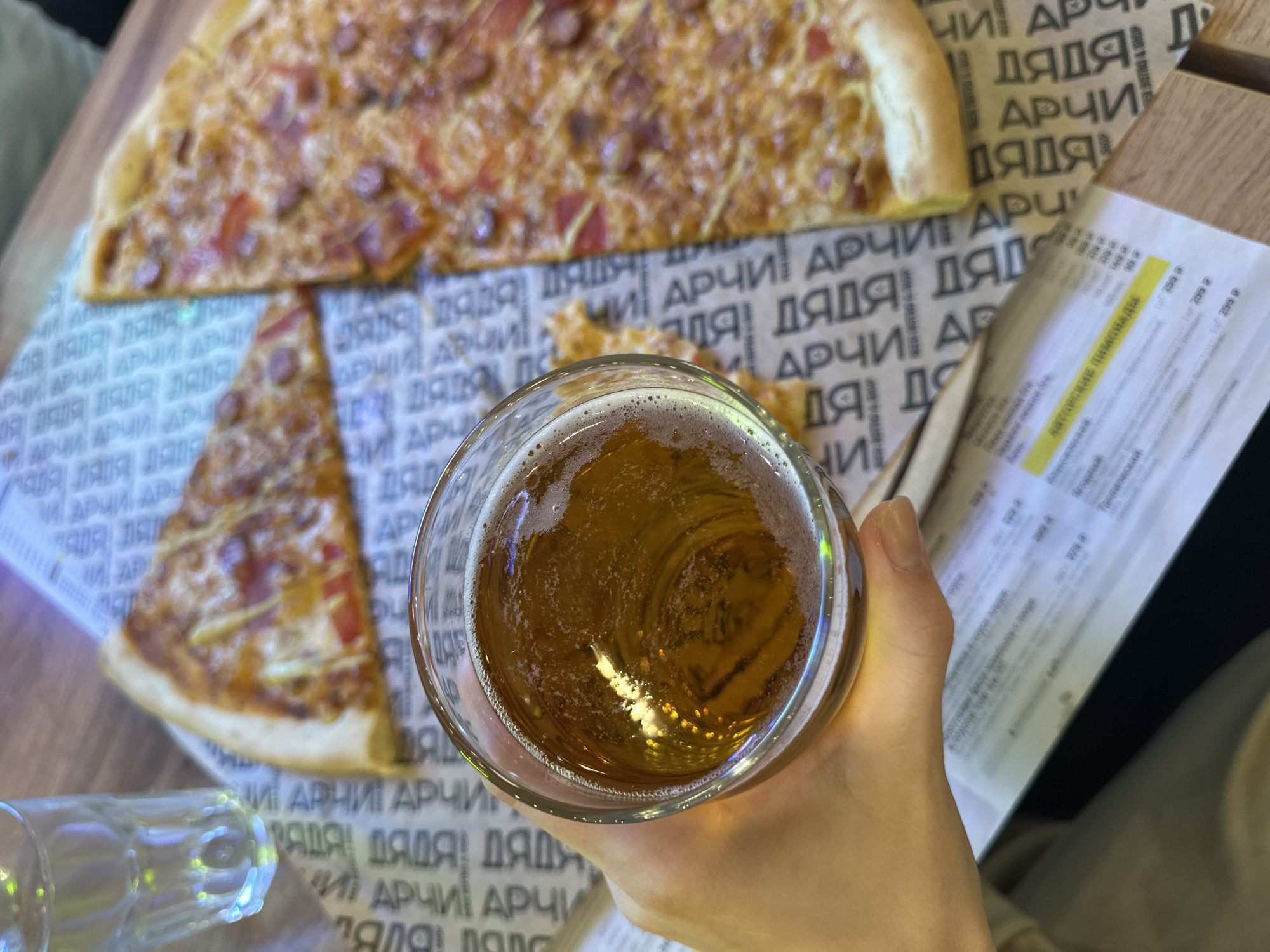 The image depicts a table scene, likely at a restaurant in Perm, Russia based on the Cyrillic text on the paper. A hand holds a glass of amber-colored beer, with light reflecting off the liquid and foam. Adjacent to the beer are slices of pizza, featuring a crust, sauce, cheese, and toppings. A menu is visible on the right, filled with text. The background is a printed paper placed under the pizza that contains the words "ДЯДЯ АРЧИ" repeated over and over. The overall setting suggests an informal meal at a restaurant.