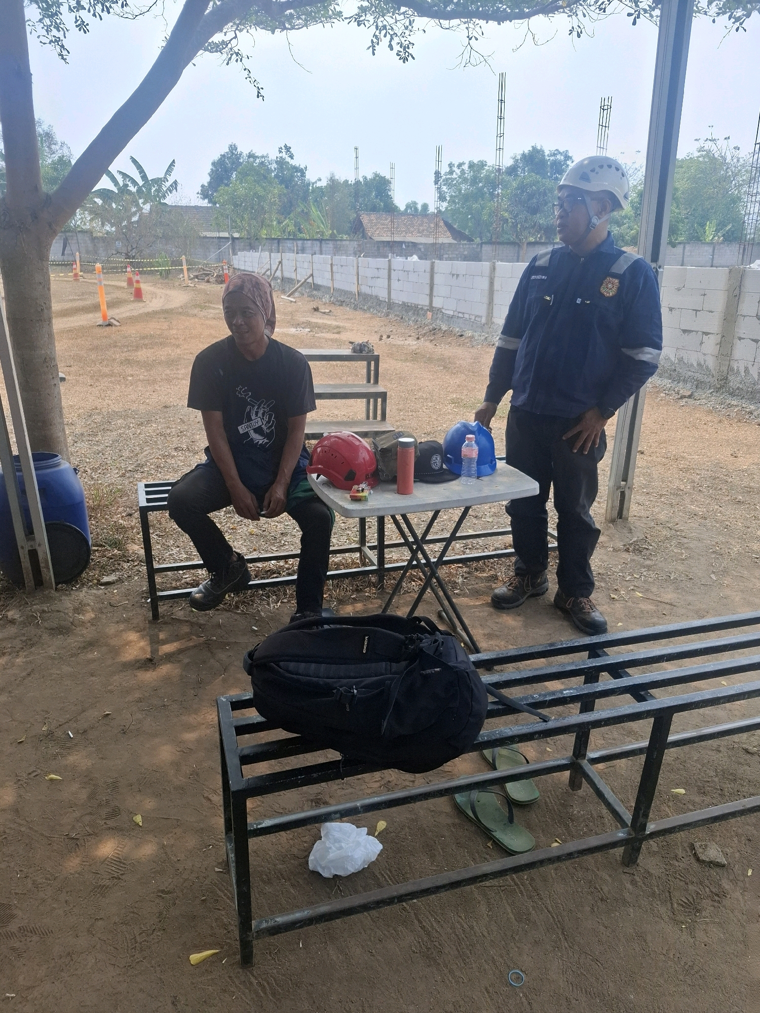In Serang, Indonesia, a construction site is depicted with two individuals. One man, wearing a dark t-shirt and headscarf, sits on a metal bench. The other man, in a blue work jacket and white hard hat, stands near a foldable table. On the table are two hard hats, a water bottle, and a thermos. A black backpack rests on another metal bench in the foreground, and a pair of green flip-flops are on the ground beside it. The ground is dirt with scattered leaves. In the background, a brick wall and construction materials are visible, along with trees and a clear sky. Orange traffic cones and yellow caution tape suggest a demarcated work area. A large blue barrel is to the left of the scene.