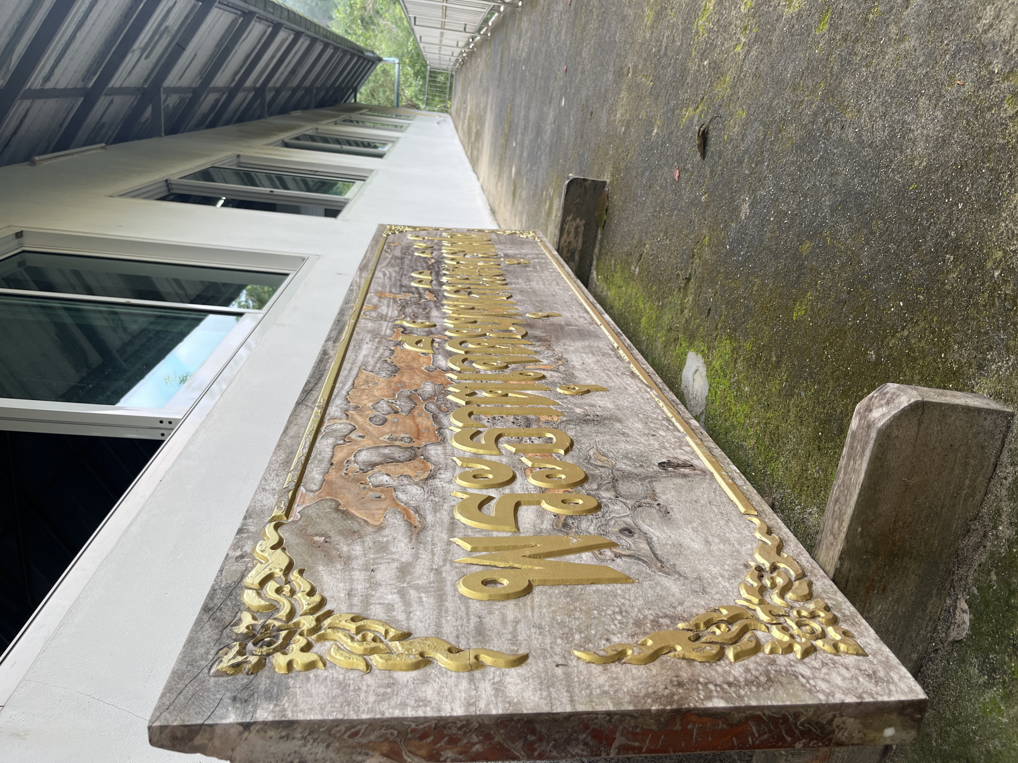 A weathered wooden sign with gold lettering and ornate gold carvings sits on a concrete walkway in Patong, Thailand. The sign is supported by two wooden blocks. The walkway is lined with a white building with several windows and a metal railing. Green moss grows on the walkway and the sign. In the distance, trees and a hill are visible. The scene appears to be outdoors, possibly in a public area.