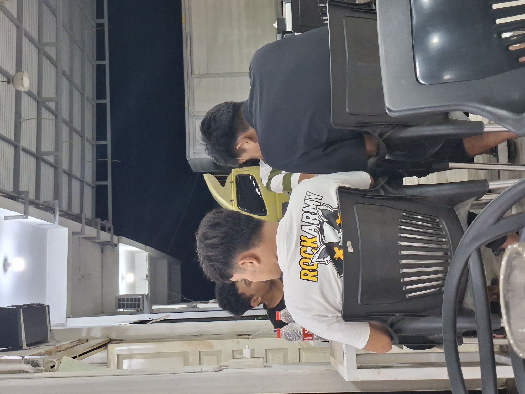 In Sigli, Indonesia, three individuals are seated at tables outdoors, under a covered area with fluorescent lighting. Two men are facing away from the camera, seated at a table in the foreground. The man on the left wears a white t-shirt with the text "ROCKARMY" printed on the back in yellow and black, alongside a graphic of a cat or wolf-like creature. He is seated in a black plastic chair with a slatted back. To his right, another man wears a black shirt and is also seated in a black plastic chair. A third individual, partially visible to the left, faces the other two and is wearing a black t-shirt. Two plastic water bottles are visible on the table in front of the man in the white shirt. In the background, a yellow truck and a white cargo truck are parked. The environment appears to be a food stall or restaurant area.