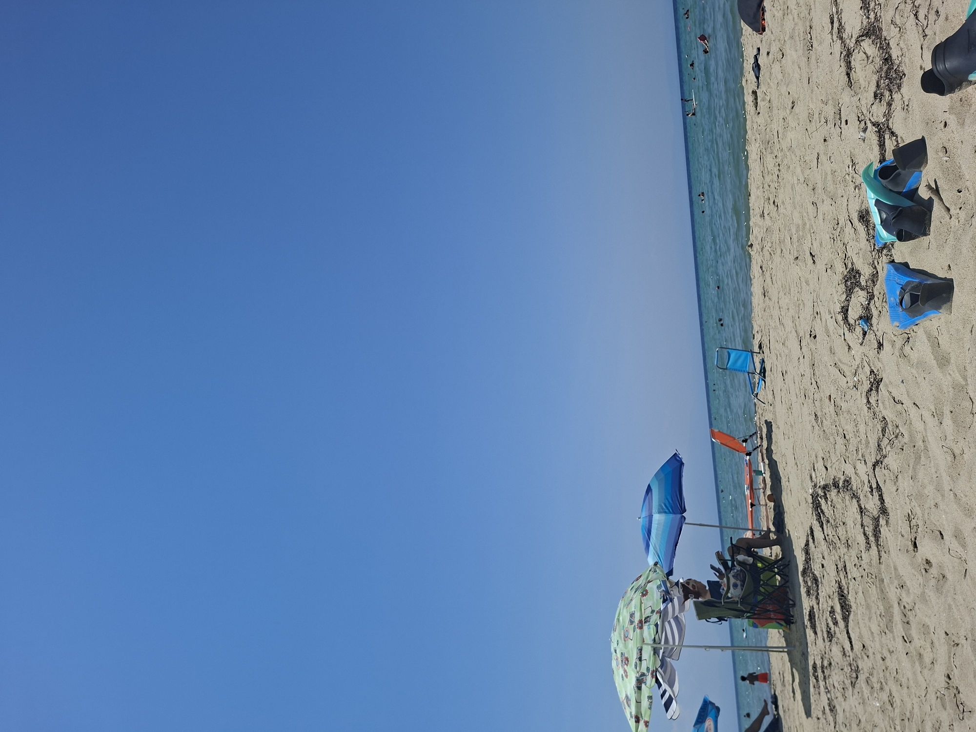 The image depicts a sunny day on a sandy beach in Nea Kallikrateia, Greece. The sky is a clear, vibrant blue. In the foreground, several pairs of flippers are laid out on the sand.
Further back on the beach, two beach umbrellas provide shade. Beneath one umbrella, a person is seated in a folding chair, appearing to read. Next to them, an orange beach chair is empty. A few other beach chairs and umbrellas are visible further down the beach.
The sea is a calm, turquoise color, with several people swimming and engaging in water activities. The shoreline is a mix of sand and some dried seaweed. The overall scene conveys a relaxed atmosphere typical of a summer day at the beach.