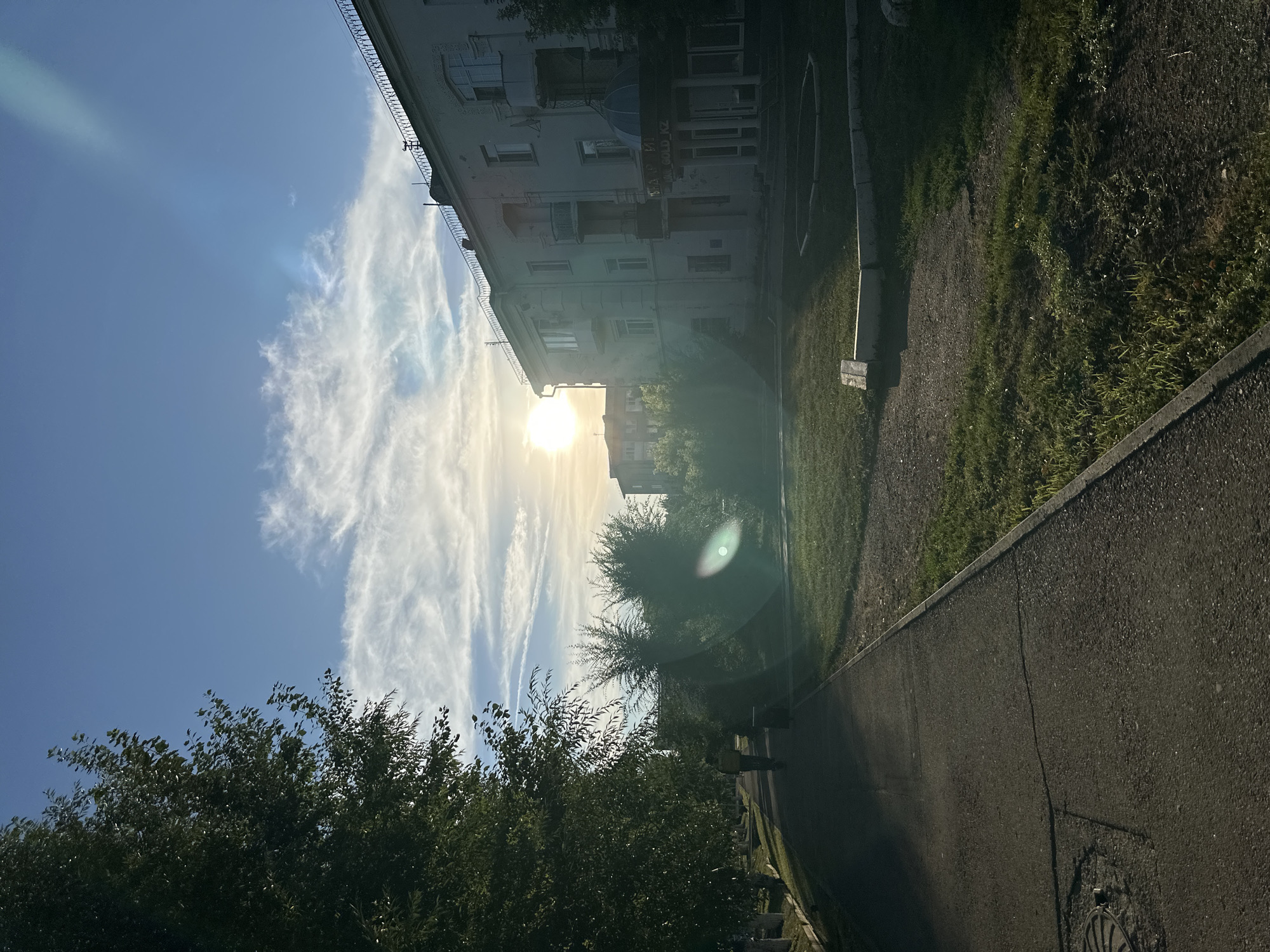 The image depicts a street scene in Kokshetau, Kazakhstan, during daylight. A paved walkway runs along the left side, bordered by a grassy area and trees. On the right, a multi-story building with balconies and windows is visible. The sun is low in the sky, casting long shadows and creating a lens flare. A lone individual wearing a yellow jacket is walking on the walkway, facing away from the camera. The sky is blue with scattered white clouds. The scene appears to be in a residential area, possibly in front of an apartment complex.