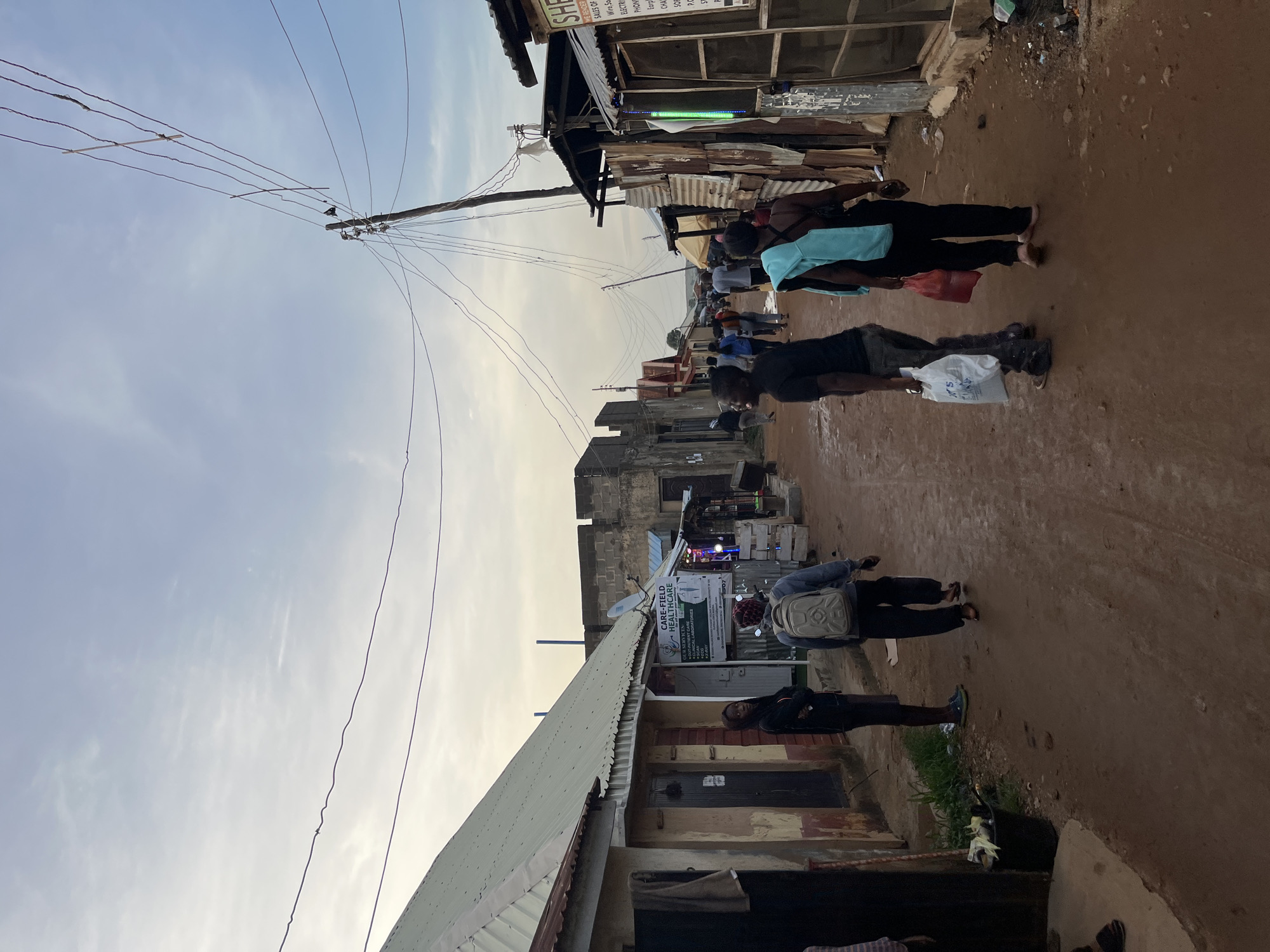 This image captures a vibrant street scene in Jos, Nigeria, depicting daily life in what appears to be a bustling, semi-urban environment. The time of day is suggested by the soft, diffused light and light blue sky with wispy clouds, possibly indicating early morning or late afternoon.
The primary subjects are several pedestrians walking along a wide, unpaved dirt road. Most are dressed casually, and some individuals are seen carrying white or red shopping bags, suggesting commercial activity or errands. The overall atmosphere appears routine and calm.
The street is lined with various structures, many of which are commercial establishments. These buildings display a mix of construction materials, including corrugated metal sheets, concrete blocks, and plaster. Overhead, numerous electrical or communication wires crisscross the sky, strung between utility poles, one of which has a bird perched on it. A satellite dish is also visible on one of the buildings.
Visible text on signs provides insight into local businesses. A prominent sign reads "CARE-FIELD HEALTHCARE," with partially visible text below it listing "OUR SERVICEN" (likely "OUR SERVICES") and "CAL LABORAT" (suggesting "Clinical Laboratory"). Another sign, partially obscured, indicates "SHE," "WE RENDER," "SALES OF," followed by items like "Wire, So" (likely sockets), "ELECTRI" (electrical), "PHON" (phones), "Earp" (earphones), and "CHA" (chargers), pointing to an electronics or general store.
The setting is characterized by the unpaved road, the functional and varied architecture of the buildings, and the general visual cues of a busy local street.
