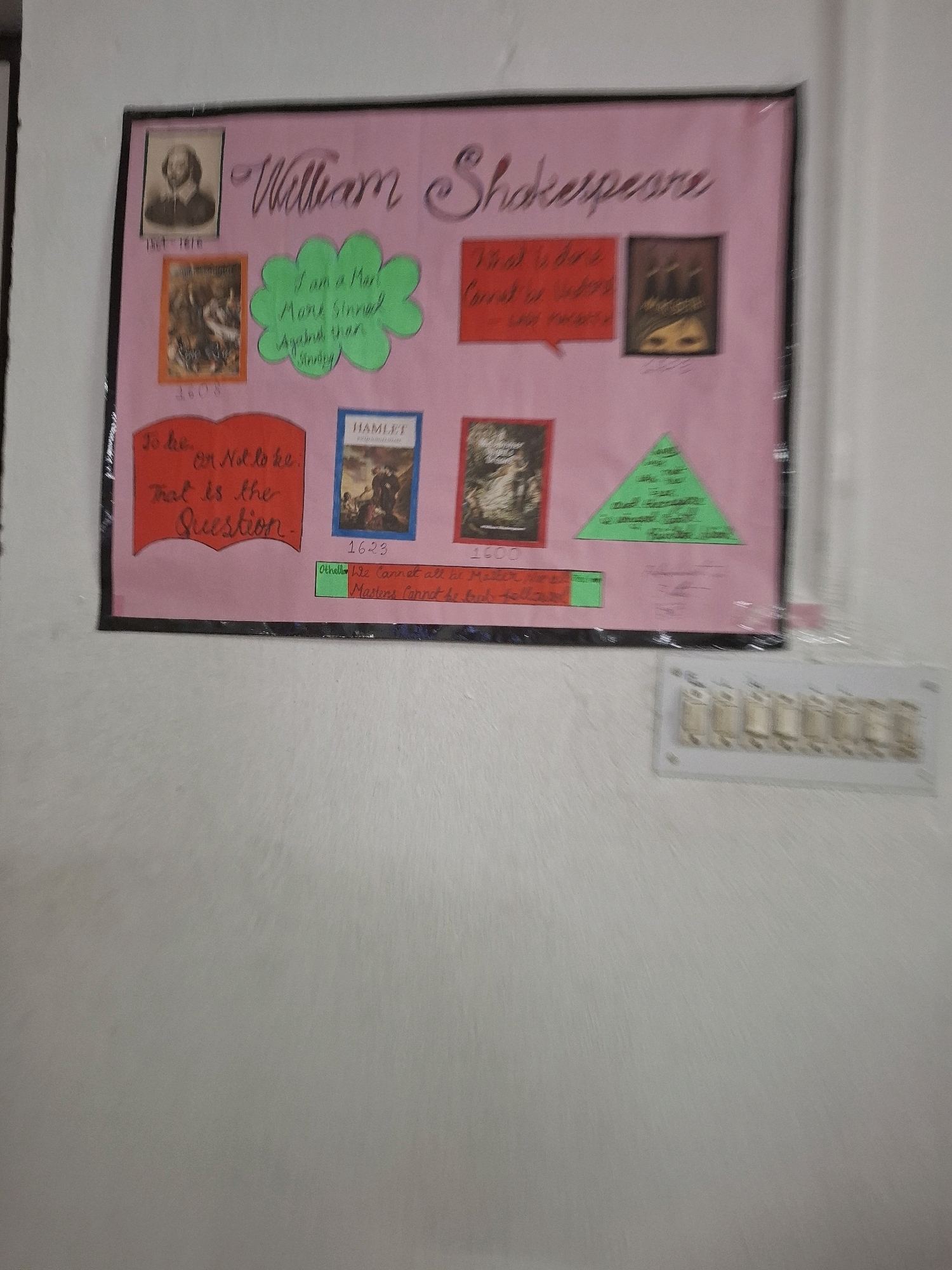 The image shows a bulletin board in Barasat, India, dedicated to William Shakespeare. The board is pink and features a portrait of Shakespeare, along with various quotes and images related to his works. There are also some handwritten notes and dates. The bulletin board is framed with black tape. In the foreground, a white electrical outlet with six switches is visible on a textured wall.