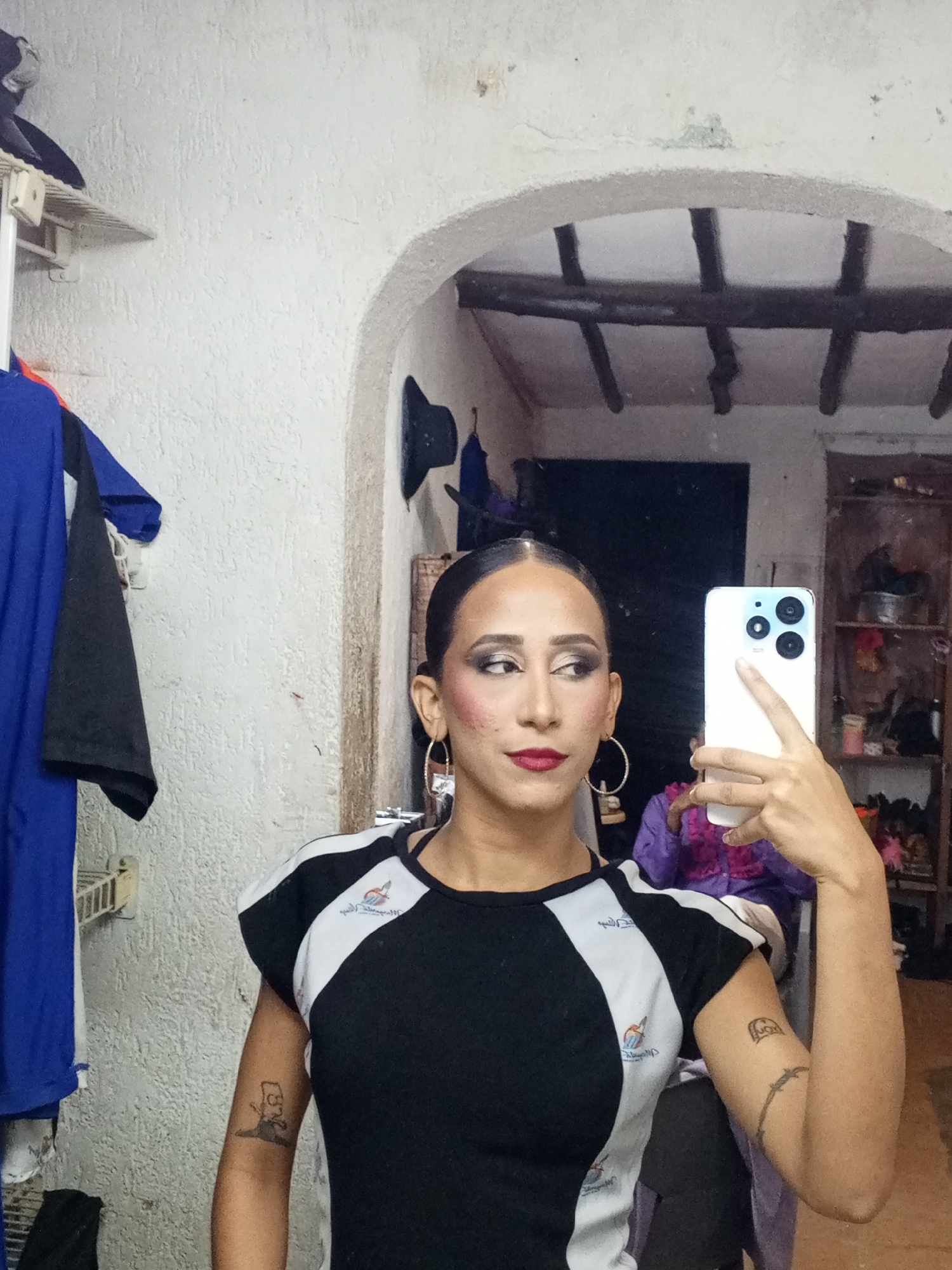A person is taking a selfie with a white smartphone in what appears to be a dressing room or backstage area. The person has dark hair pulled back, hoop earrings, and is wearing makeup including dark eyeshadow and red lipstick. They are wearing a black and white athletic-style top. The right arm of the person is visible, revealing a tattoo of a barbed wire and a tattoo of Bart Simpson.
Behind the person, a mirror reflects part of the room, including a ceiling with wooden beams and a wooden shelf unit with various items. To the left of the person, there is a clothing rack with several garments hanging, including a blue shirt. The walls are white and have a textured appearance.
Based on the appearance of the person and setting, the image seems to have been taken indoors, possibly in a private or backstage area in the city of Porlamar, Venezuela.