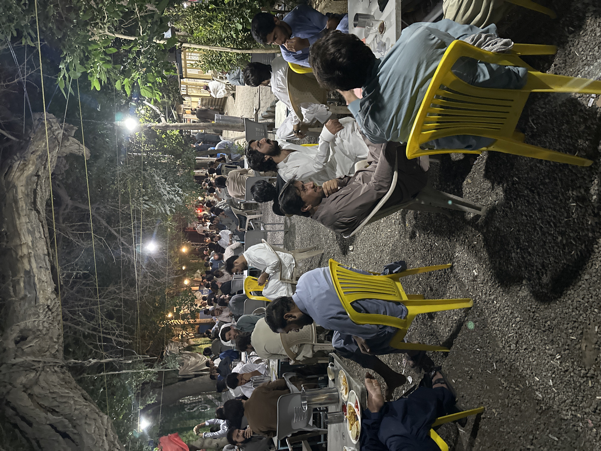 A large group of men are seated outdoors at tables. The location is identified as Quetta City Tehsil, Pakistan, by a previous prompt. The men are diverse, dressed in traditional Pakistani attire, including shalwar kameez in white, blue, and brown hues. They are seated in yellow and gray plastic chairs at tables with plates, cups, and other items. Several men are eating, conversing, and relaxing. The outdoor setting has gravel ground, trees, and string lights. The scene is illuminated by overhead lighting.