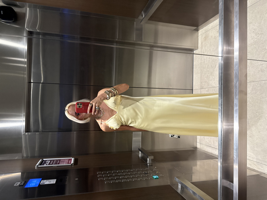 A person with short, platinum blonde hair, wearing a long, light yellow slip dress, pink-tinted sunglasses, layered necklaces, and numerous stacked bracelets on their left wrist, is captured in a selfie. The individual holds a red smartphone, obscuring their face, while a small white or silver bag is held in their left hand. The scene is set inside a modern elevator, characterized by brushed stainless steel walls and doors, with a dark wood panel accent on the right wall. The left wall features an elevator control panel with multiple buttons, a light-up green button, and a digital display indicating "S2-S1". A vertical information sign is mounted above the control panel. Polished metal handrails are present on the left wall and reflected at the bottom of the frame. The floor consists of light-colored, square tiles. This upscale elevator environment is consistent with high-end hotels, residential towers, or commercial complexes found in Dubai, United Arab Emirates.
