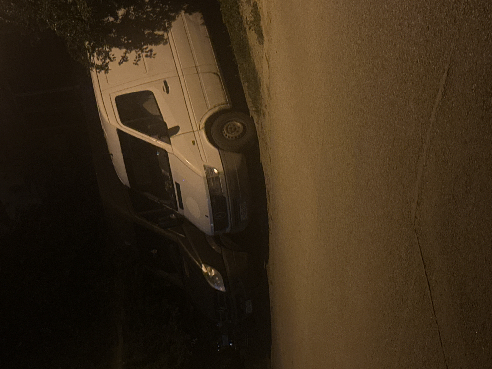 The image depicts a dimly lit outdoor scene, likely at night, with two vehicles parked on a street. The foreground shows a cracked asphalt surface. The primary focus is a white Mercedes-Benz Sprinter van parked on the right side of the frame, angled slightly upwards from left to right. The van has its headlights on, casting a faint glow. To its left, a darker vehicle, possibly a black Mercedes-Benz sedan, is parked. The background is obscured by darkness and foliage on the right, suggesting the presence of trees or bushes. The scene appears to be in a residential or semi-urban area. Based on the available information about the city of Sliven, Bulgaria, this image could represent a street scene within the city.
