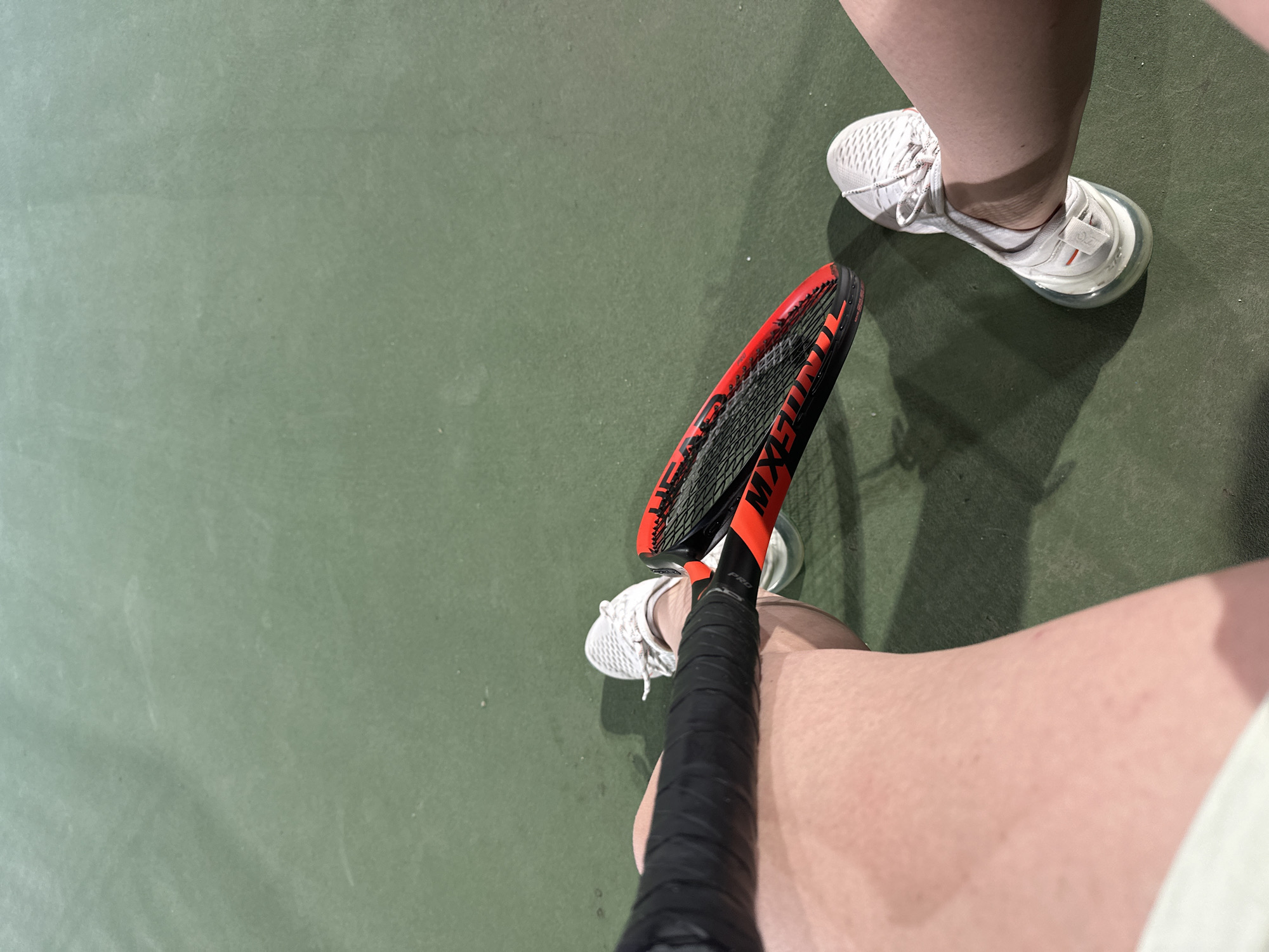 The image shows a person's legs and a tennis racket on a green tennis court. The person is wearing white athletic shoes. The tennis racket is black with red accents. The court is outdoors, and the lighting suggests it may be late afternoon or early evening. The specific location within the city of Sofia, Bulgaria, cannot be determined from the image.