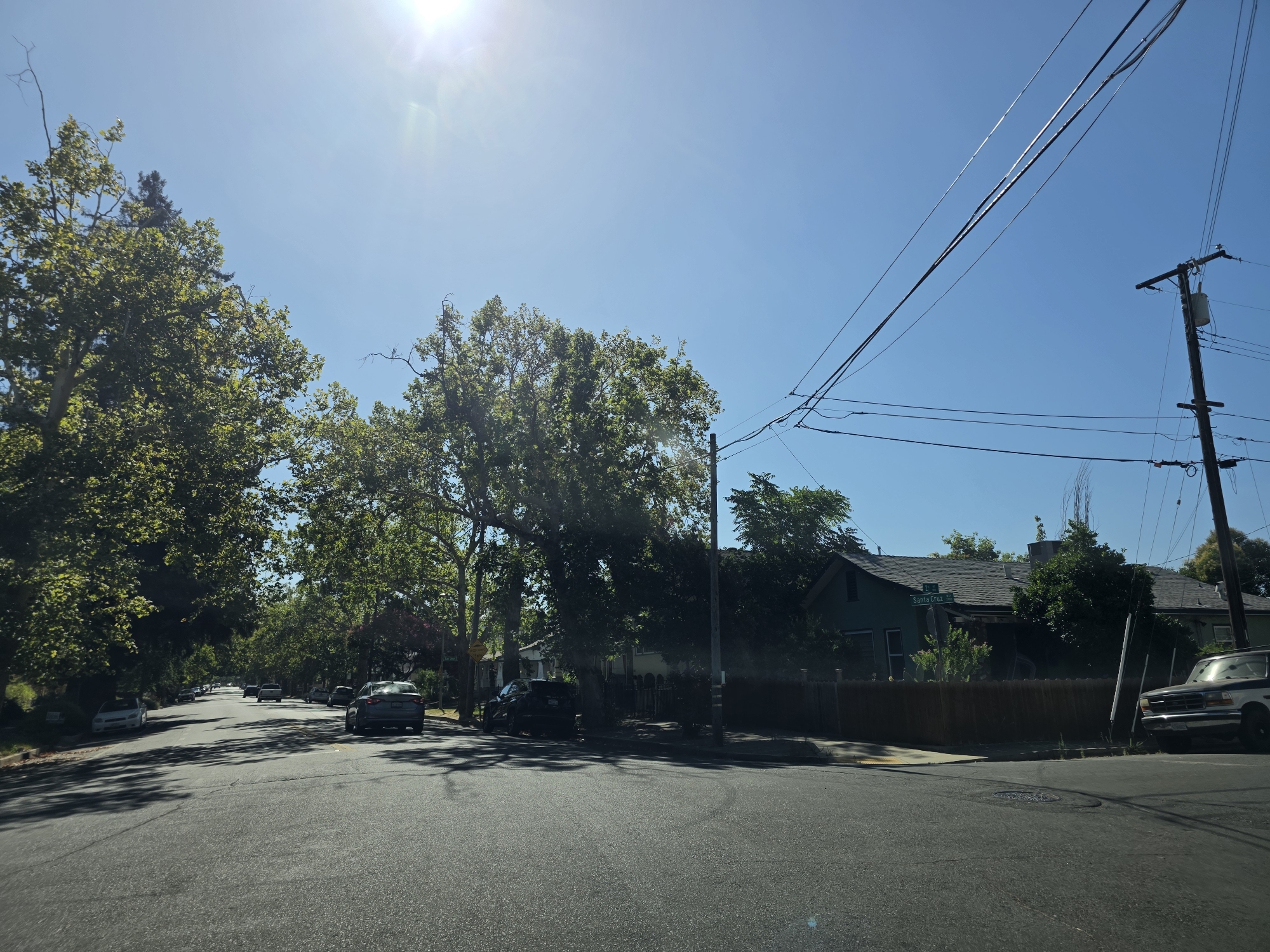 This media file depicts a residential street scene in Sacramento, California, under a bright, sunny sky. The time of day appears to be late morning or early afternoon, with the sun high and casting distinct shadows. The weather is clear and warm, indicated by the bright sunlight and the presence of lush green trees lining the street.
The scene is dominated by a two-lane asphalt road with several cars parked along the sides and a few in motion. The architecture of the houses visible suggests a suburban setting, with some single-story homes and a mix of architectural styles. A wooden fence runs along the property line in front of one of the houses. Overhead, power lines stretch across the sky, supported by a wooden utility pole.
One notable detail is a street sign at an intersection, clearly visible and reading "Santa Cruz Ave". The presence of mature trees with dense foliage suggests that this is a well-established neighborhood. The overall mood is peaceful and ordinary, capturing a typical day in a residential area. There are no people visible in the frame.