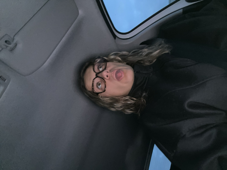 A person with curly blonde hair, wearing glasses and a black coat, sits in a car. The individual is looking up at the camera with their tongue sticking out. The car's interior is visible, including the roof, sun visor, and window. The sky is visible through the window. The image is taken from a low angle, looking upwards at the subject.