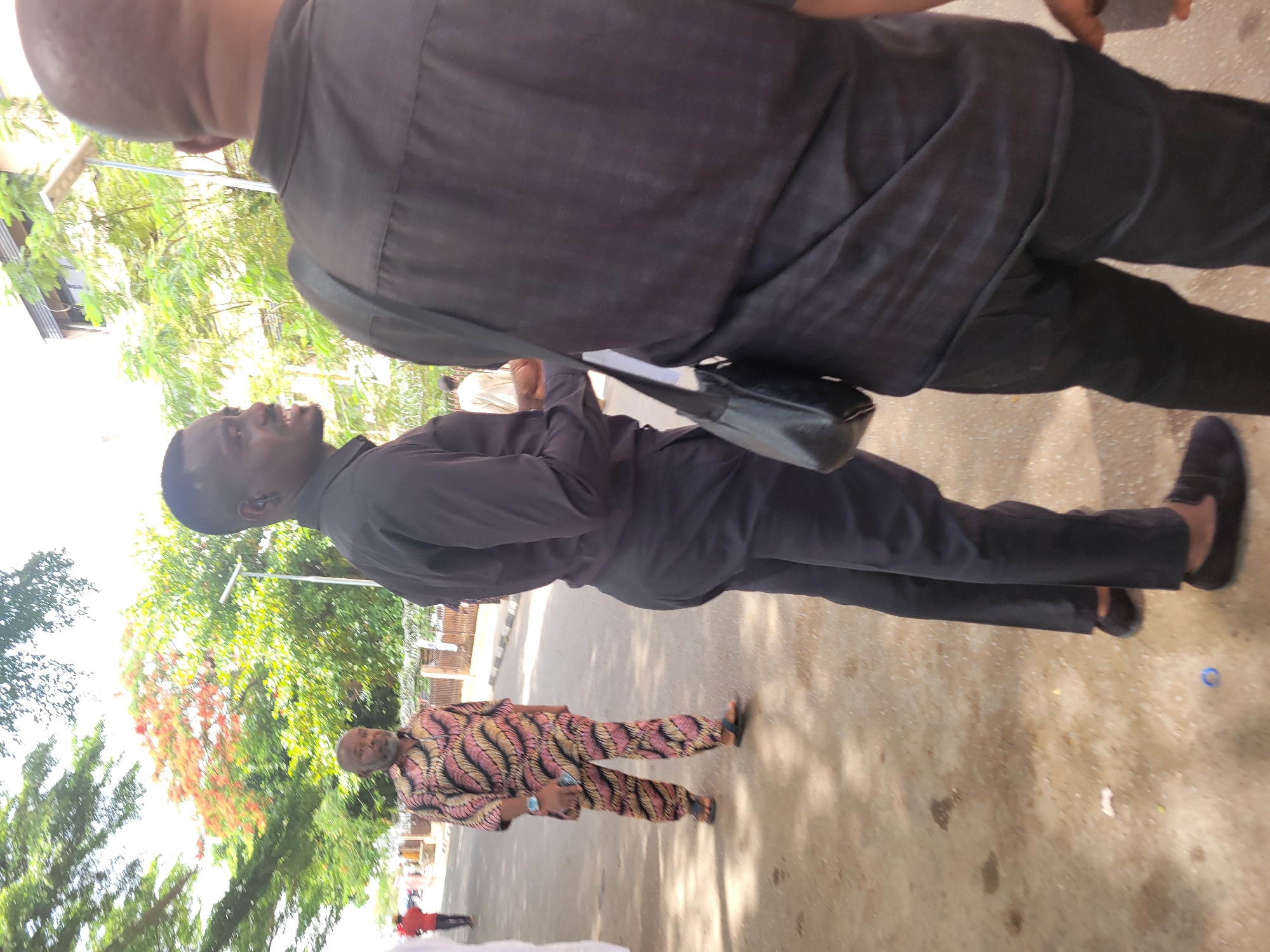 The image captures an outdoor scene, likely a street or pathway in Abuja, Nigeria, under bright daylight. Three main figures are visible, along with indistinct background elements.
In the immediate foreground, from a slightly overhead perspective, is the back of a person's head and shoulders, wearing a dark (likely black) collared shirt.
In the middle ground, a man stands, angled slightly to his right, wearing a black long-sleeved shirt and black trousers, paired with dark slip-on shoes. He has a small black shoulder bag slung across his body. He holds what appears to be papers or a small notebook in his left hand and is looking towards the left side of the frame with a clear smile on his face, indicating engagement or amusement. He has a short beard.
Further in the background, another man with a grey beard is visible. He is wearing a colorful, traditional patterned long-sleeved shirt and matching trousers, with sandals on his feet. He is looking generally towards the viewer's right, possibly towards the person in the middle ground.
The ground is paved with concrete or asphalt, showing some scattered debris, including a blue circular object and a white piece. The area is lined with lush green trees, some displaying reddish-orange foliage, characteristic of a tropical environment. The bright lighting suggests it is daytime, and the clear skies indicate sunny weather. Several other indistinct figures can be seen in the distant background, partially obscured by trees and sunlight. No legible text is visible in the image.