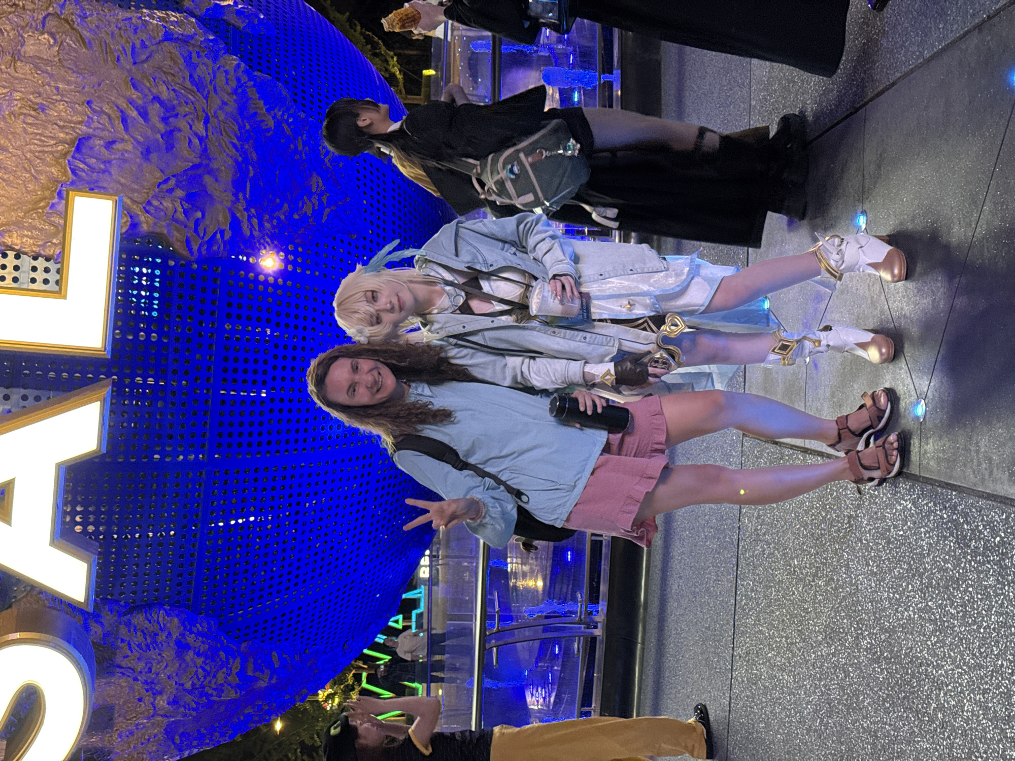 The image shows a group of people standing in front of a large, illuminated blue globe. The globe features a textured surface and is partly obscured by large, white letters. One person is cosplaying as a female anime character with blonde hair and is wearing a white and blue outfit with gold accents, complete with white and gold boots. Another person stands beside them wearing a light blue jacket, pink shorts, and sandals. They are holding a water bottle and making a peace sign. Other people are visible in the background. The location could be a theme park or entertainment venue. The city of Tongzhou District, China, cannot be confirmed from visual evidence alone.