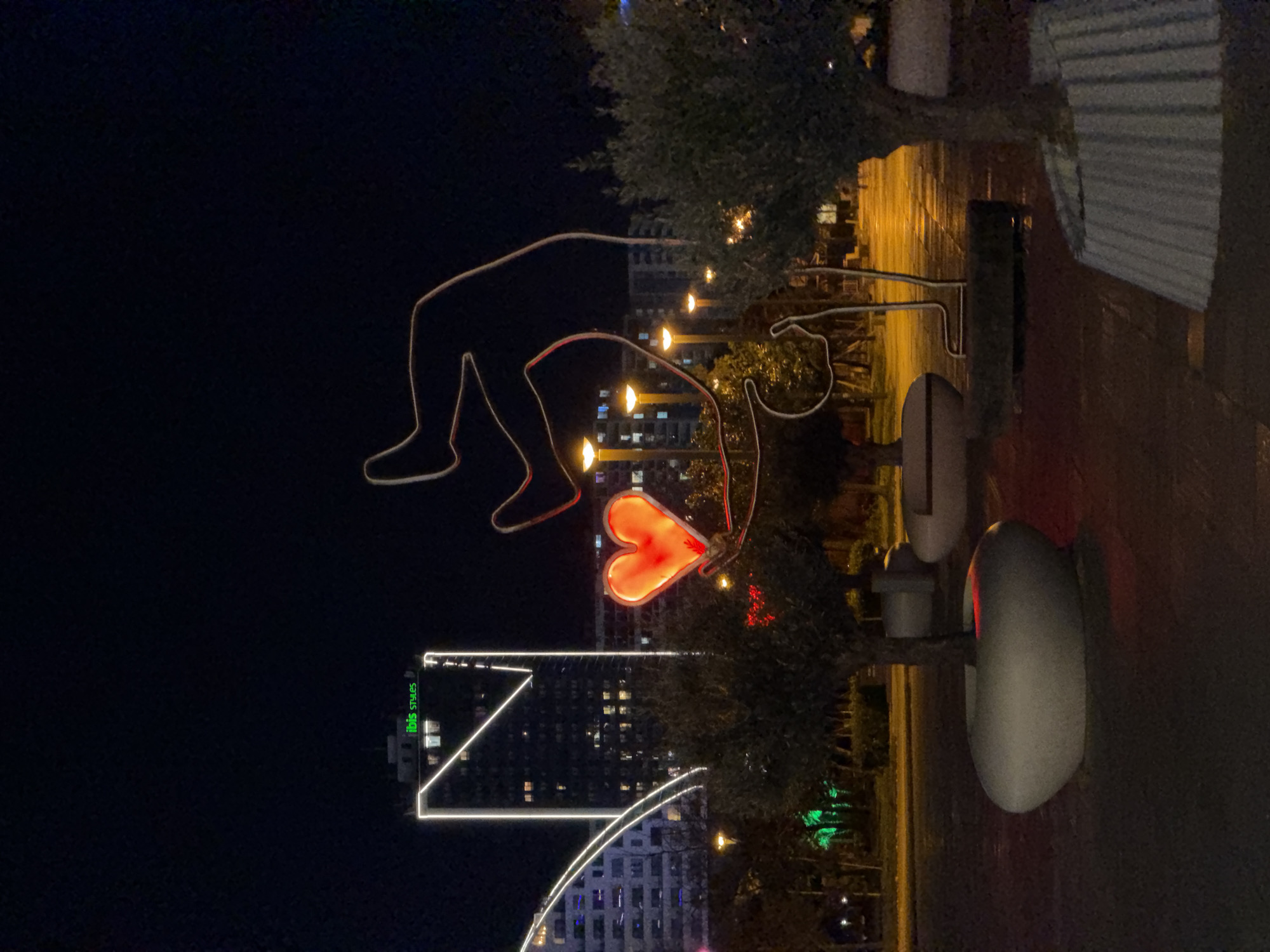 At night in Batumi, Georgia, a brightly lit sculpture of a man, bent over in a yogic pose with a heart shaped outline, faces the Ibis Styles hotel, which has a brightly lit outline. The scene is partly obscured by foliage. A small bus stop is visible on the right of the image.