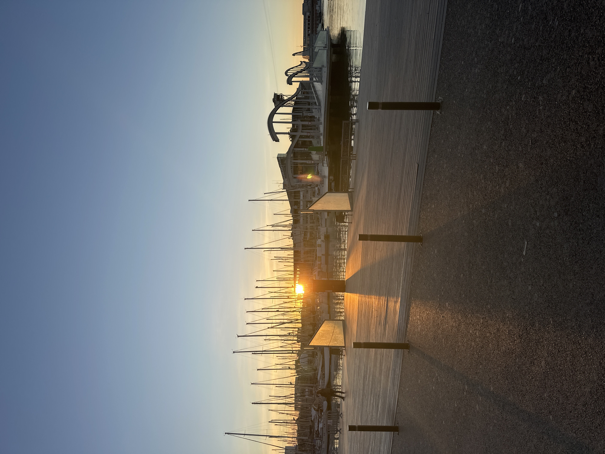 The image depicts a marina at sunset in Barcelona, Spain. Many sailboat masts are silhouetted against the setting sun. A wooden boardwalk with benches is visible in the foreground. The Rambla de Mar bridge can be seen in the background.