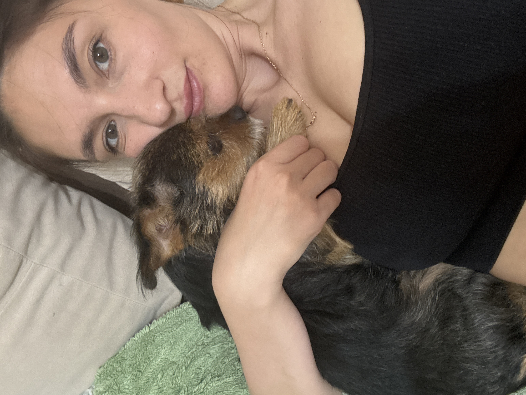 A woman is lying on a bed, cuddling a brown and black dog. The woman has light skin, brown hair, and brown eyes. She is wearing a black tank top and a gold necklace. The dog is snuggled against her chest, and she is holding the dog with one hand. The bed has a light-colored pillow and a green towel on it. The location is unknown and cannot be determined from the image.