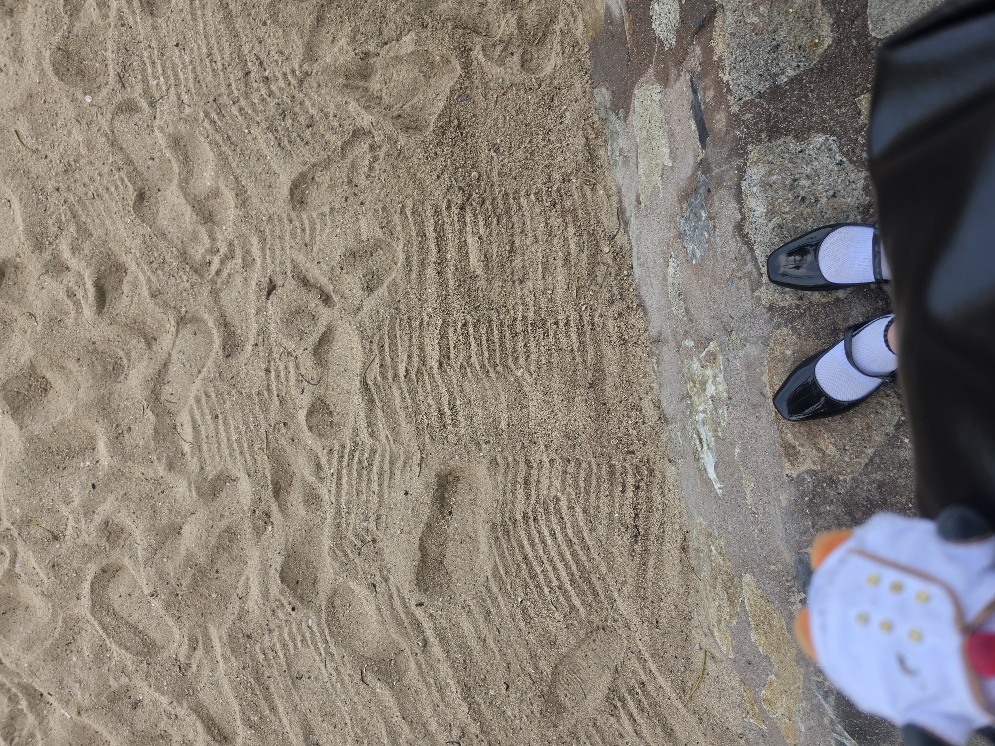 The image shows a patch of sand and a stone pathway with a pair of black patent leather shoes and white socks. The shoes and socks are partially obscured by a black object, possibly a bag. A white object with orange accents is also visible. The location is Pattaya City, Thailand. The sand has various textures, including footprints and parallel lines.