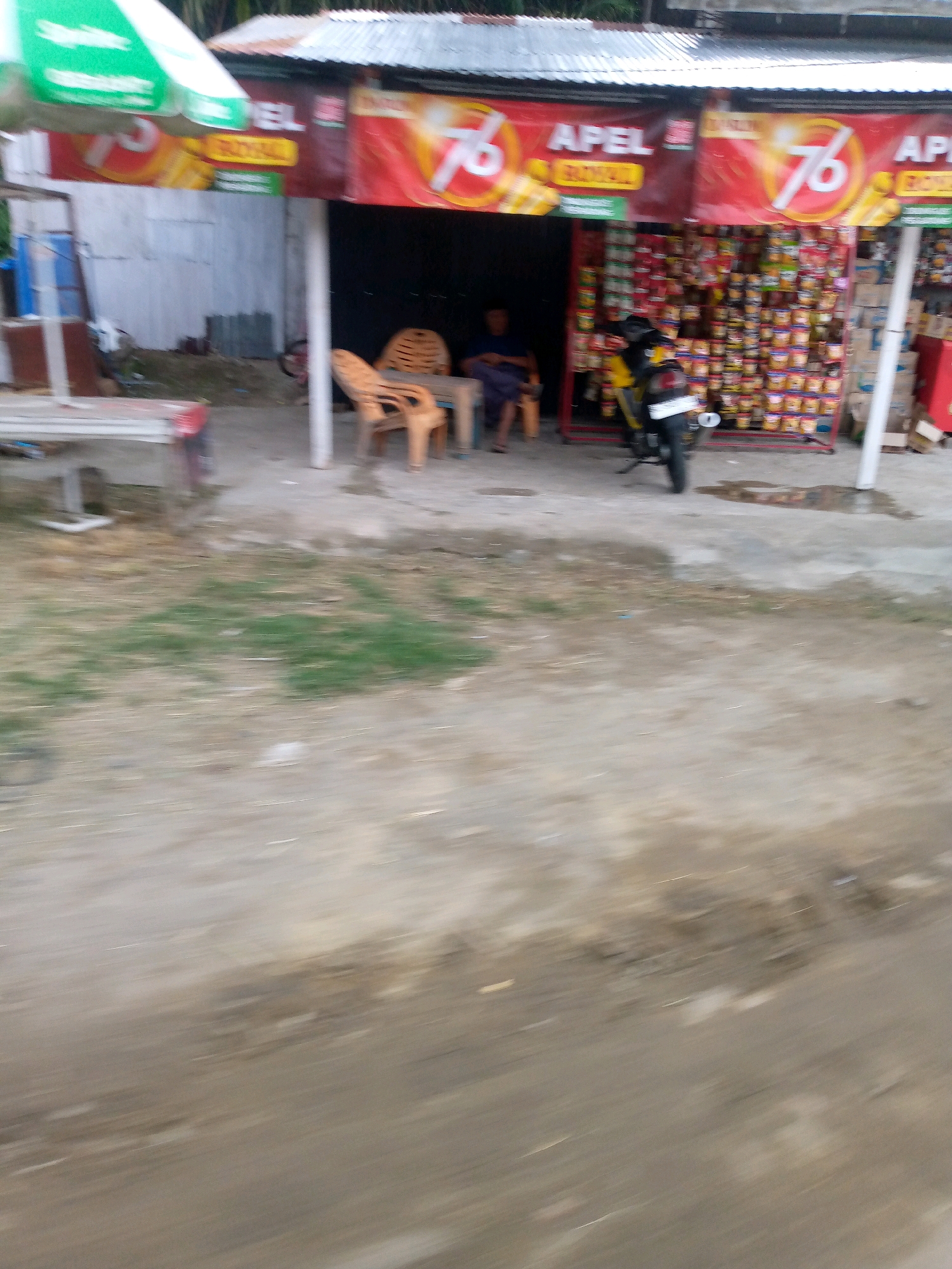 The image depicts a roadside scene in Cibrektunong, Indonesia, with a small shop in the background. A corrugated metal roof covers the shop, which displays red banners with yellow text and the number "76". The shop appears to be selling various products, likely snacks or groceries, stacked on shelves. In front of the shop, there is a motorcycle parked, and two chairs are visible, one occupied by a person wearing a dark shirt and blue pants. A small table is also present. The foreground of the image is a dirt and gravel path, with some patches of grass. The overall image is blurry, suggesting it was taken while in motion.