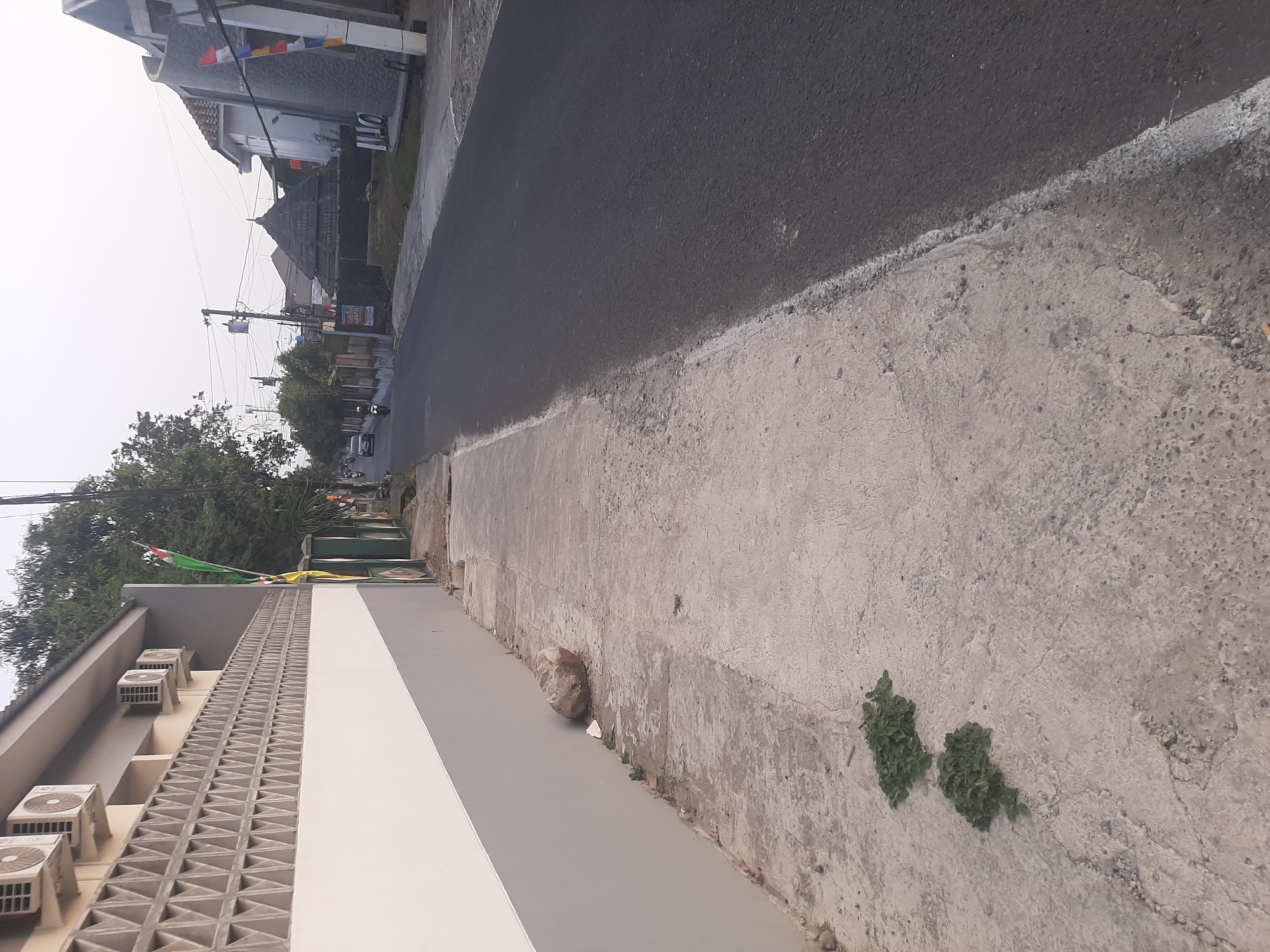 This image captures a street scene in Sumampir, Indonesia, on a bright, overcast day. On the left, a building with a light gray and white facade features a decorative wall of triangular vents and several air conditioning units mounted on the upper section. A sidewalk runs alongside the building, made of cracked concrete with patches of green weeds. A large, rounded stone rests on the sidewalk near the wall, along with some white debris. Further down the sidewalk, two clusters of green weeds are growing through cracks in the concrete.
The street itself is paved with dark asphalt and curves gently to the right. On the right side of the street, there are houses with various architectural styles. One house has a dark, conical roof and a stone facade. Another appears to be under construction with a curved, dark stone exterior. Flags of red and white are visible on some of the buildings and poles. Trees line the street, and power lines crisscross overhead. A few vehicles, including a white car and a motorcycle, are seen on the road in the distance. The sky is a pale, hazy white, indicating diffused sunlight. The overall impression is of a quiet residential or semi-commercial street.