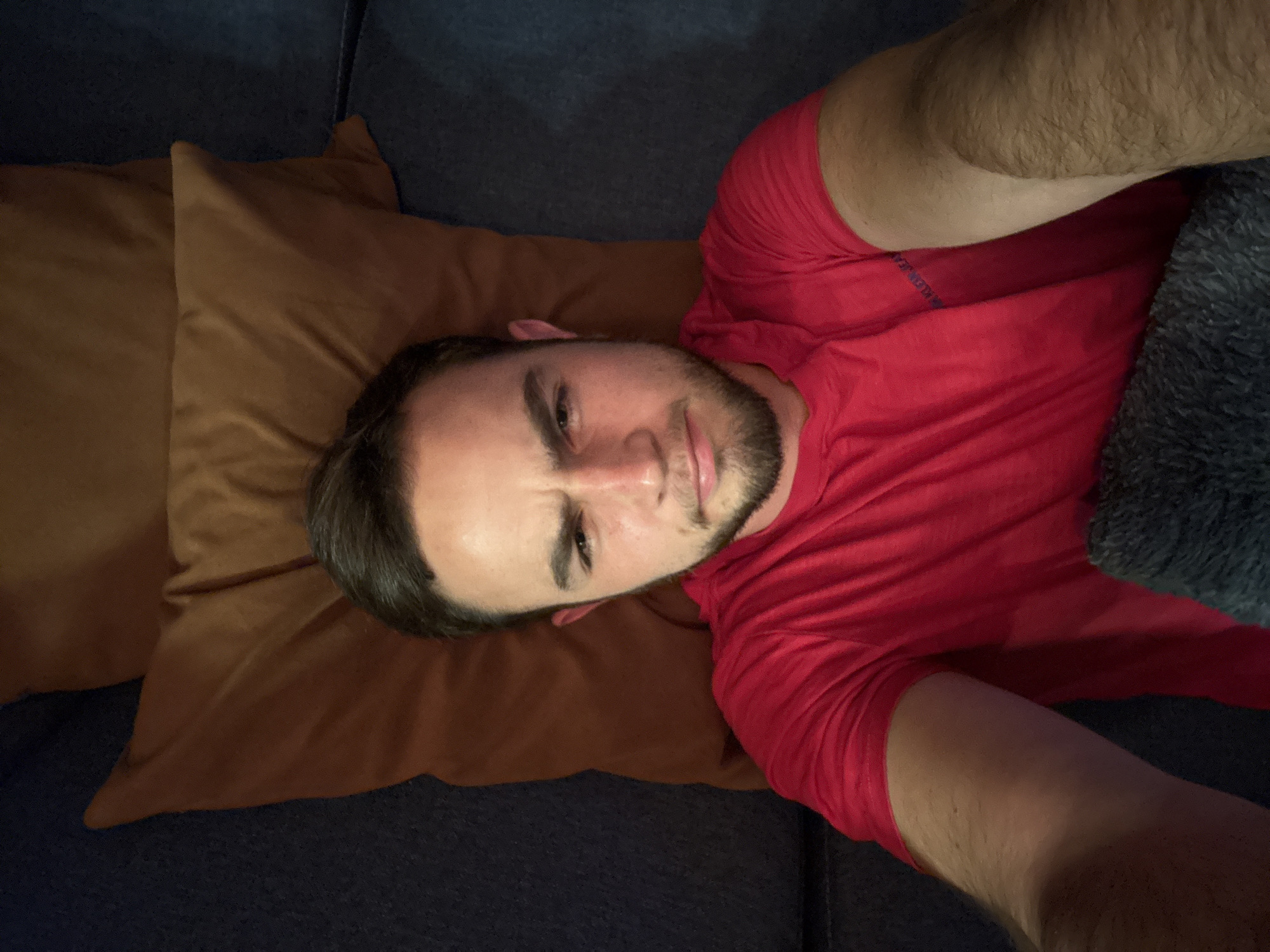 A man with short dark hair and a trimmed beard lies on a dark blue sofa. He is wearing a red t-shirt and partially covered by a grey blanket. His head rests on a brown pillow. The scene is located indoors, likely in a private residence. The city of San Bernardino Tlaxcalancingo, Mexico is not discernible from the image.