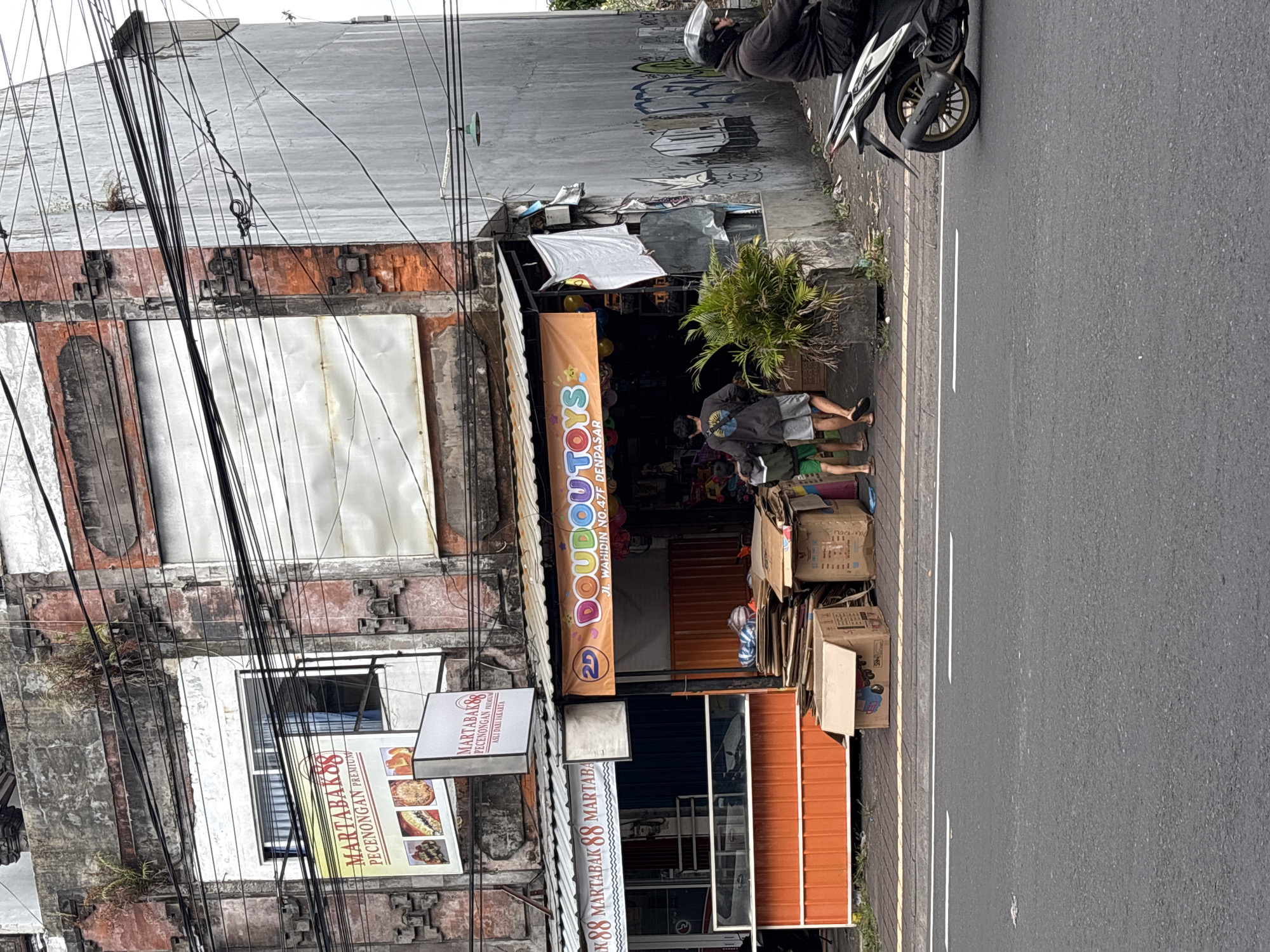 The image shows a street scene in Denpasar, Indonesia. On the left, there is a shop with an orange facade and a sign that says "MARTABAK 88 MARTABAK". Above this shop, there is another sign that advertises "MARTABAK 88 PECENONGAN PREMIUM ASLI DARI JAKARTA". To the right of the martabak shop is a toy store with a banner that reads "DOUBOU TOYS JL. WAHIDIN NO.47F DENPASAR". In front of the toy store, there are several stacks of cardboard boxes. Two young boys are standing near the entrance of the toy store, and a man is walking away from the camera, with another man on a motorbike passing by on the street. The buildings in the background appear weathered and are covered in a network of electrical wires. The street is paved with asphalt.