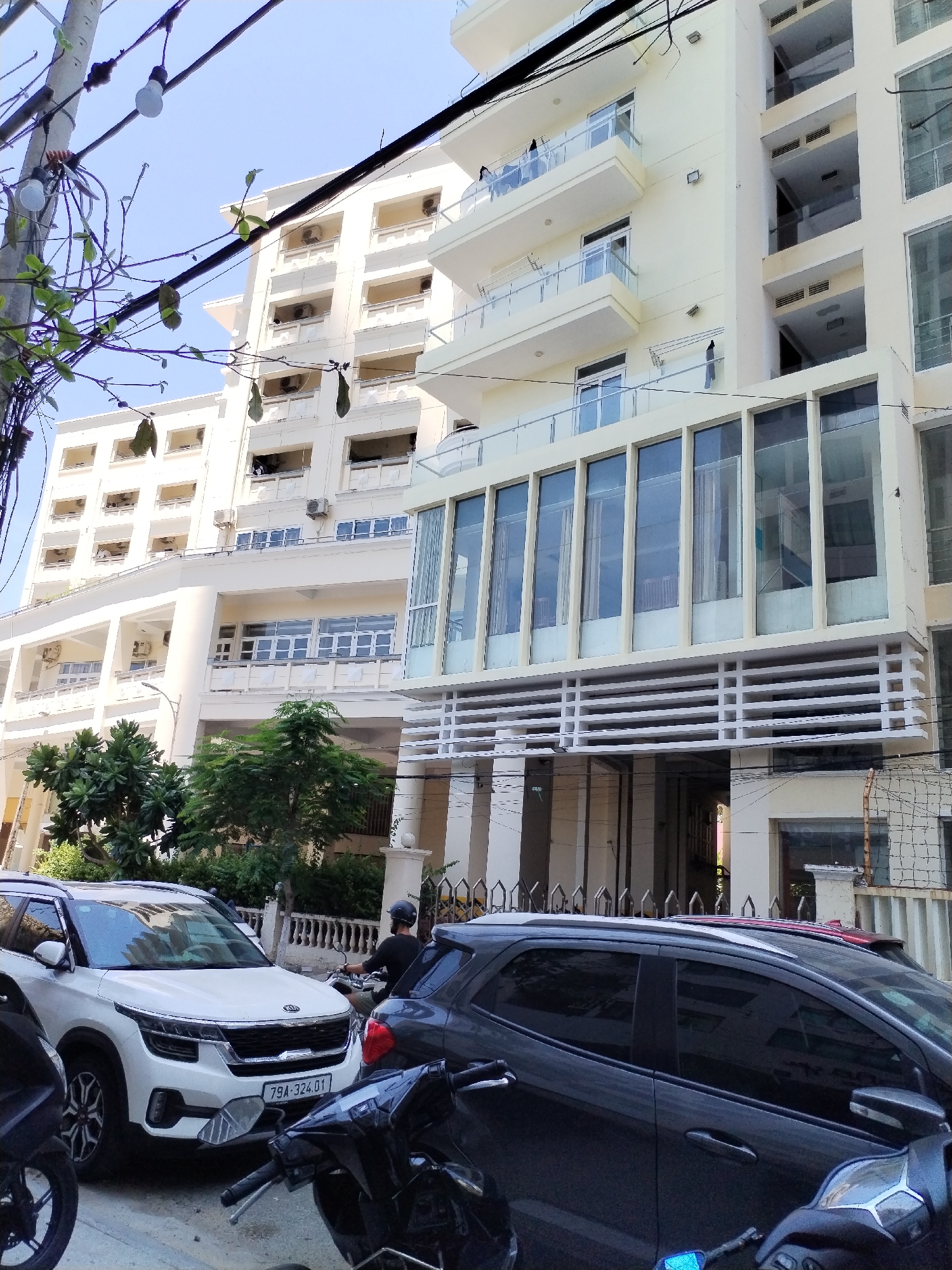 The image shows a street scene in Bac Nha Trang Ward, Vietnam, with a modern apartment building as the dominant feature. The building is light yellow and has multiple floors with balconies and large glass windows. In the foreground, there are several vehicles parked, including a white Kia Seltos SUV with license plate "79A-324.01" and a dark-colored SUV. A person wearing a helmet is seen riding a motorcycle. Trees and power lines are also visible, suggesting an urban environment. The sky is clear and blue.