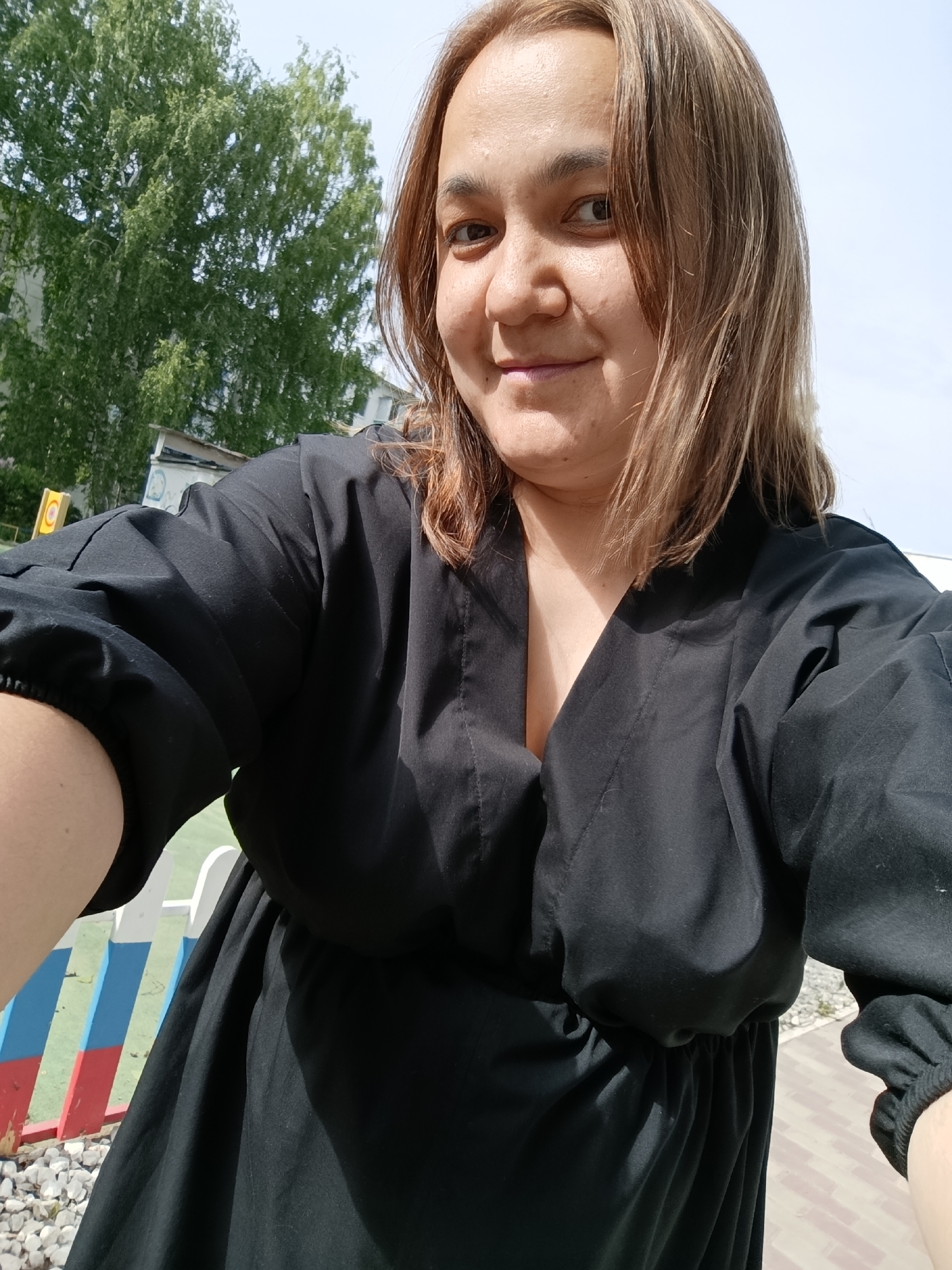 A woman is taking a selfie outdoors. She is wearing a black dress. Behind her is a tree and a building. The location is Chaikovsky, Russia, as evidenced by the Russian flag painted on a structure near the woman. The woman is on a paved walkway, and there are rocks on the ground next to her.