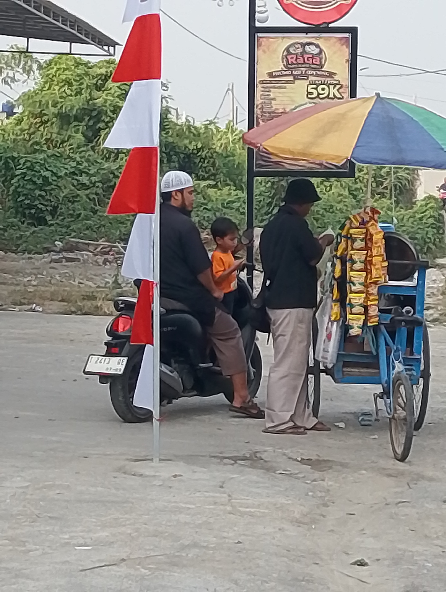 In the city of Karawang, Indonesia, on what appears to be a street or roadside, a man wearing a white skullcap and a dark shirt is seated on a black motorcycle. Next to him, a child in an orange shirt is also seated on the motorcycle. Another man, wearing a black hat and a light-colored long-sleeved shirt and pants, stands beside a blue pushcart laden with packaged goods. A colorful umbrella is attached to the pushcart. Red and white flags are displayed vertically, with one flag attached to a pole near the motorcycle. A sign with the text "RAGA" and "59K" is visible behind the umbrella. The ground is paved with a mixture of concrete and dirt. Lush green foliage forms the background.