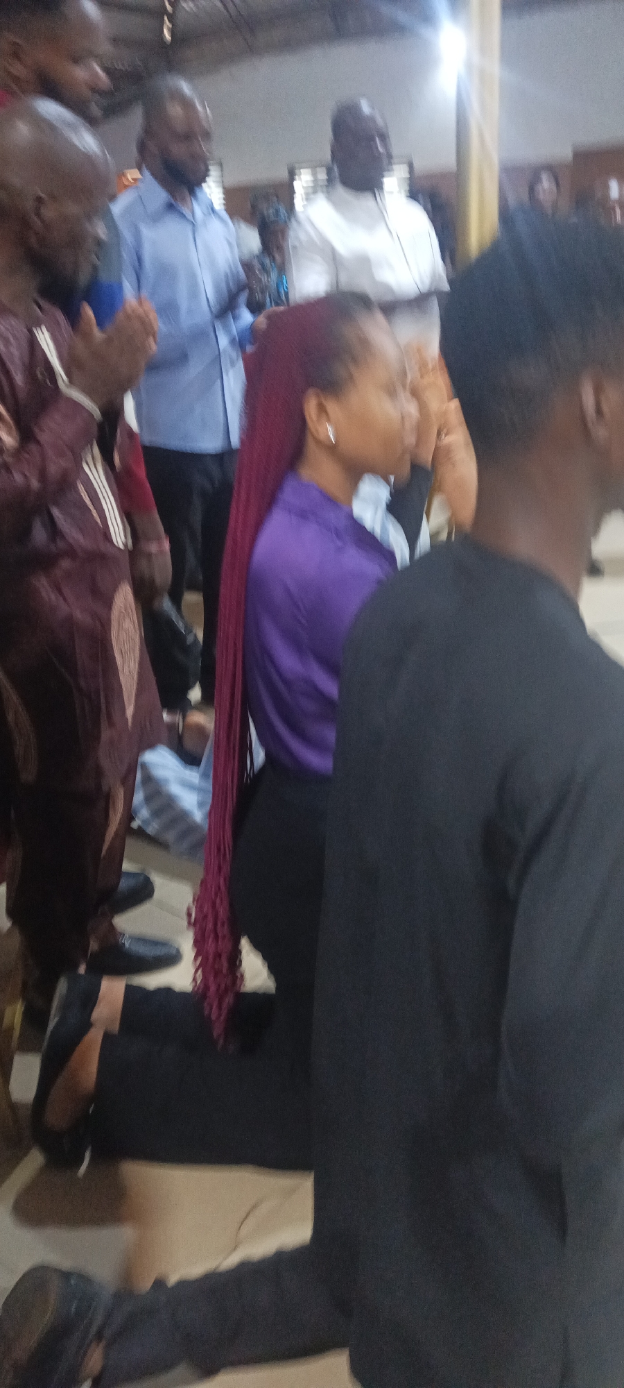 The image depicts a group of people in what appears to be a religious setting in Rukuba Road, Nigeria. Several individuals are kneeling, possibly in prayer. A woman with long, red-colored braids is kneeling in the foreground, wearing a purple blouse and black pants. To her right is a man in a black shirt. To her left, there is another man wearing a brown traditional outfit and black shoes. Other individuals, some wearing blue and white shirts, are standing behind them. The lighting is somewhat dim and the overall setting seems to be indoors.