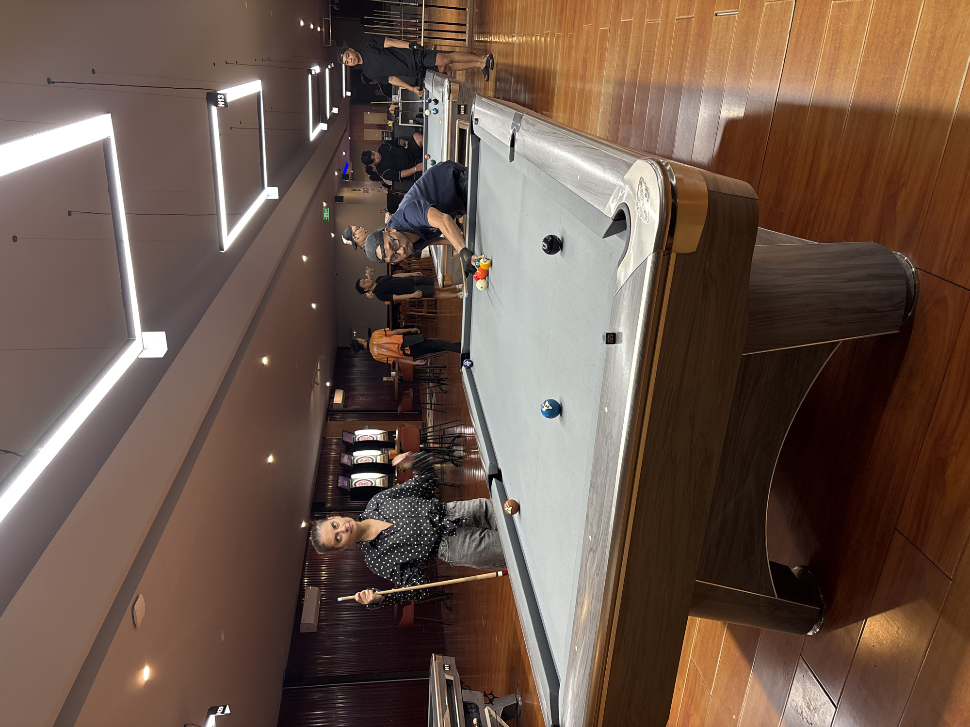 In Kuta Selatan, Indonesia, a group of people are engaged in a game of billiards within an indoor entertainment venue. The scene is illuminated by modern, linear ceiling lights. In the foreground, a woman stands near a billiard table, holding a cue stick and looking towards the camera. On the table, several billiard balls are visible, including a brown and white spotted ball and a blue ball. Another billiard table is in the mid-ground where a man wearing glasses and a cap is actively playing, positioned to strike a cluster of colorful balls. Several other individuals are also present, some standing or observing the game, contributing to a lively atmosphere. The environment features polished wooden floors and dark, paneled walls in the background, with additional dartboards visible.