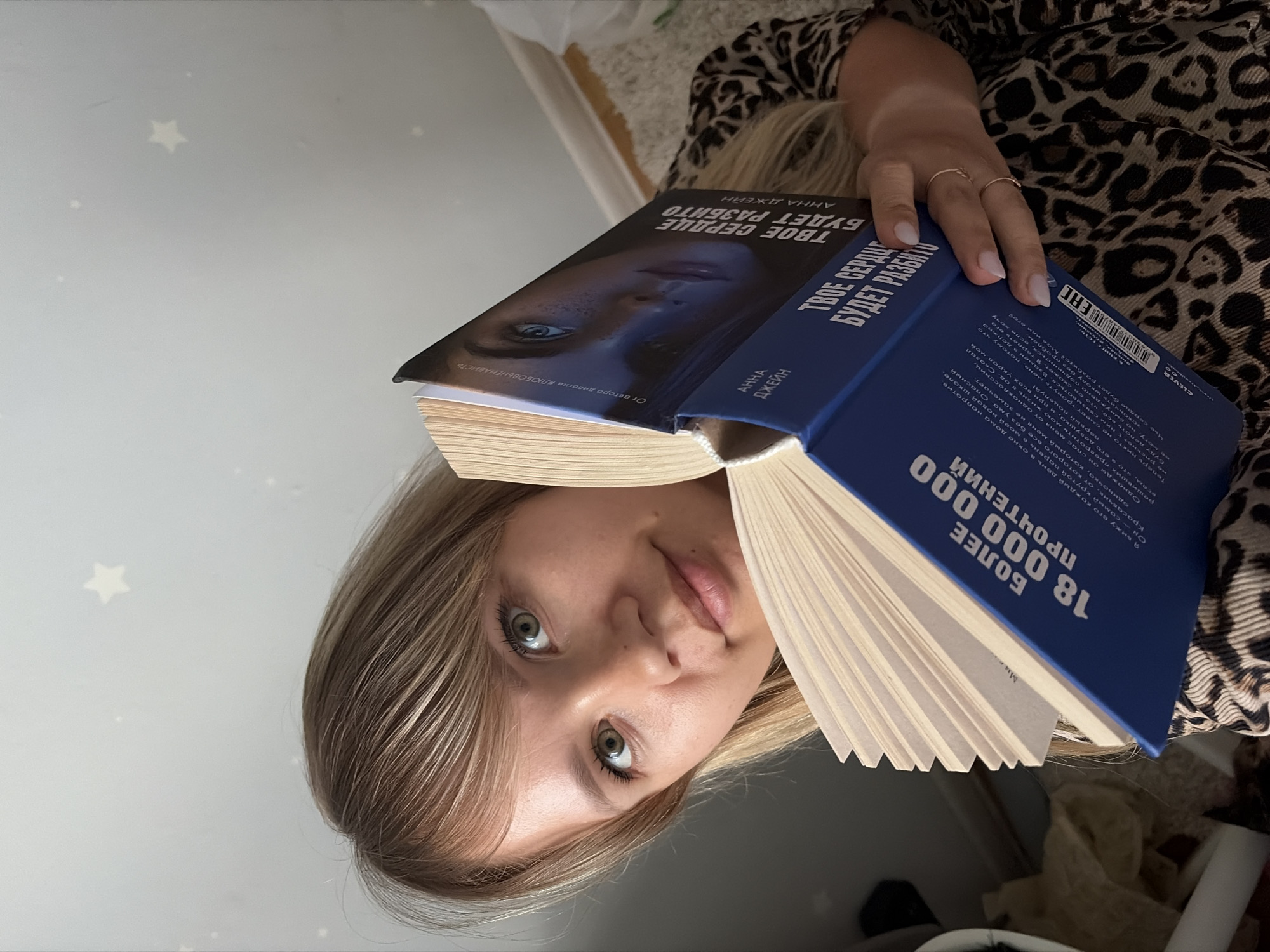 A young woman with blonde hair and green eyes is sitting indoors, holding an open book. She is looking directly at the camera, with the book partially obscuring her face. The book is blue and has text and an image of a woman's face on the cover. The text on the book is in Russian. The background appears to be a wall with star-shaped decorations. The woman is wearing a leopard-print top. There are no recognizable landmarks or details that would suggest a specific location in Chaikovsky, Russia. The image is a close-up shot, focusing on the woman and the book.