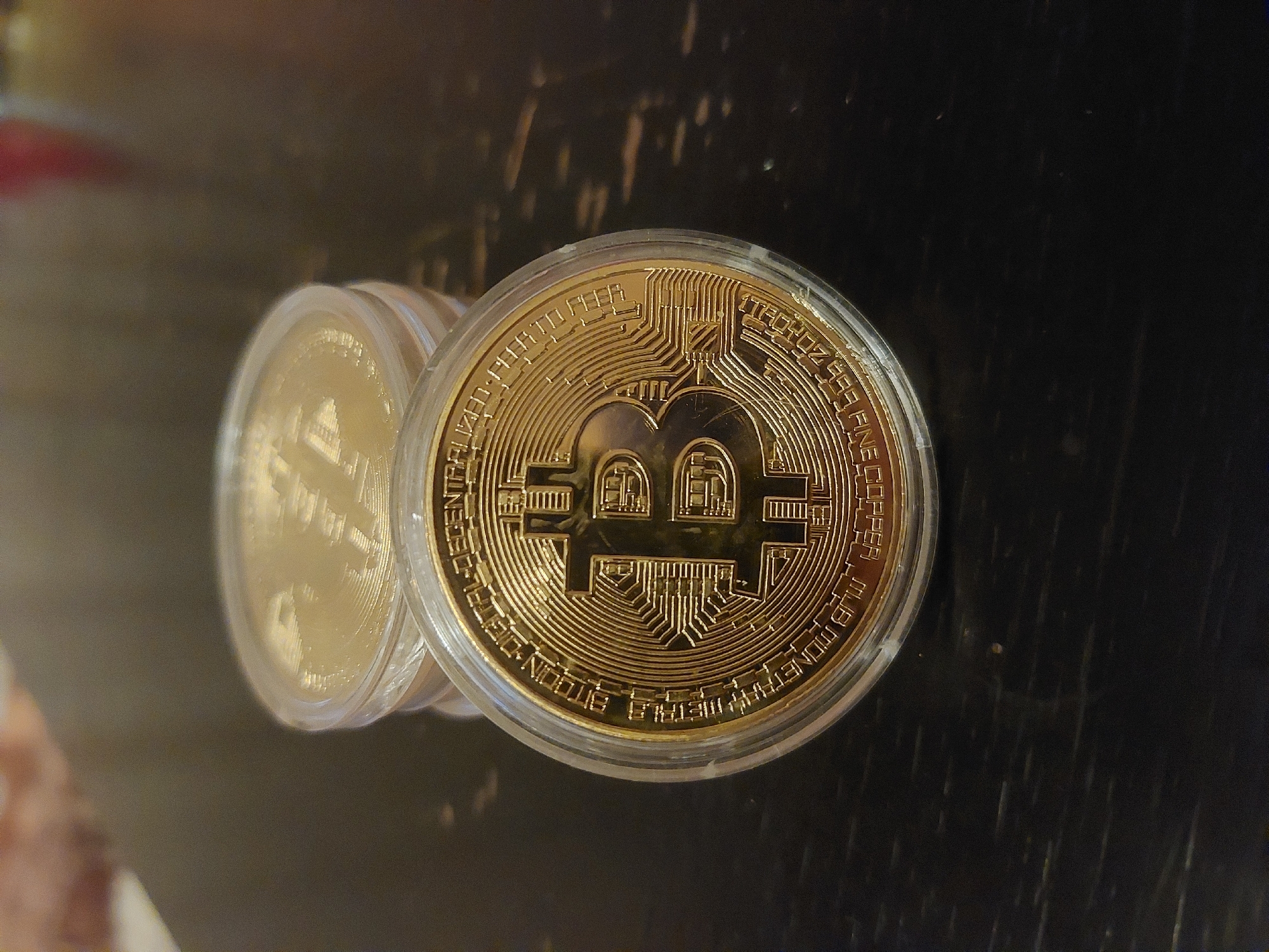 The image shows two gold-colored Bitcoin commemorative coins stacked on a dark, possibly wooden, surface. The coin in the foreground is in focus, revealing the Bitcoin logo and intricate circuit pattern design. Both coins appear to be encased in clear plastic protectors. The background is out of focus. There is no information in the image that identifies the location of the picture.